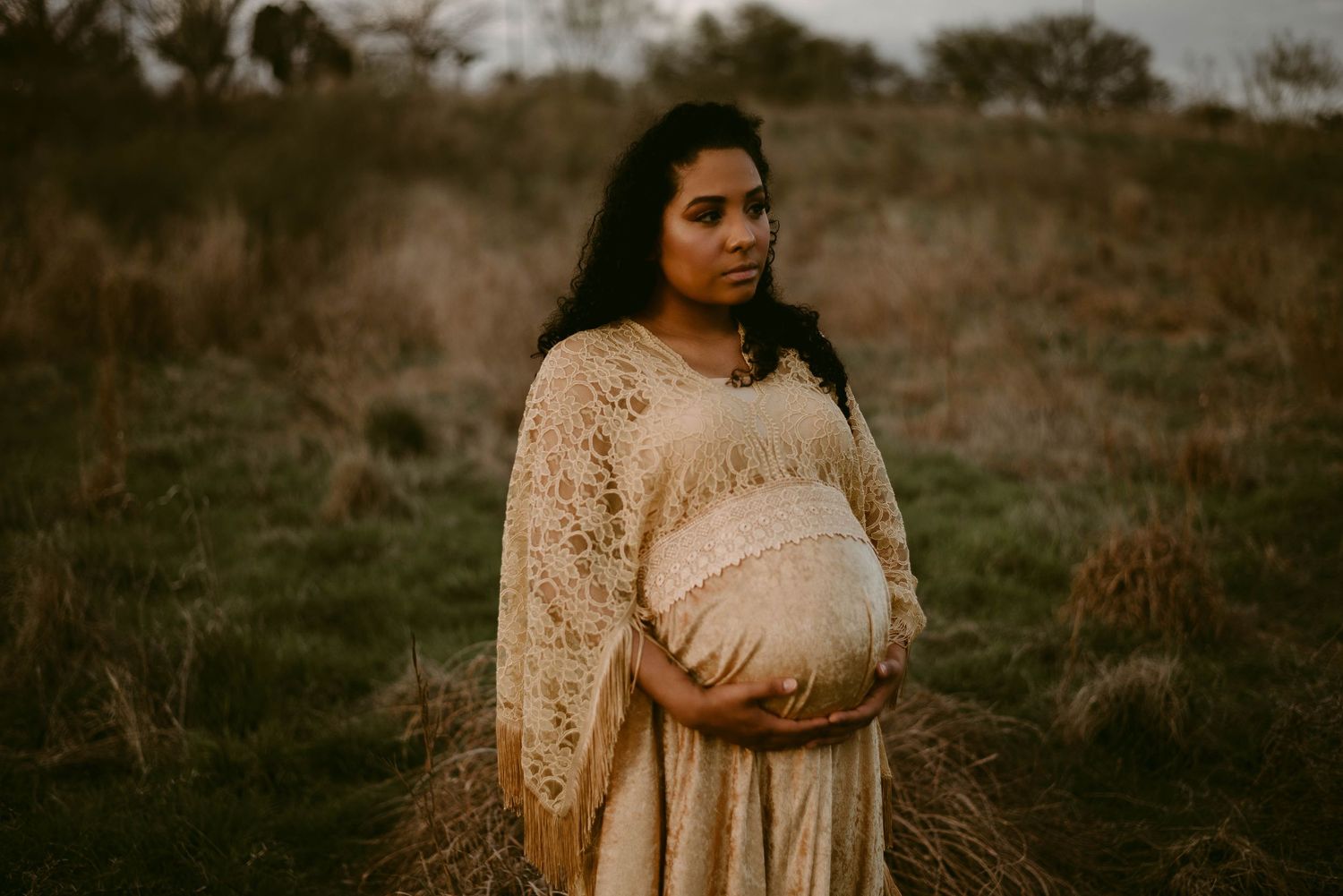 A moody maternity portrait featuring soft cream lace against a dusky outdoor landscape.