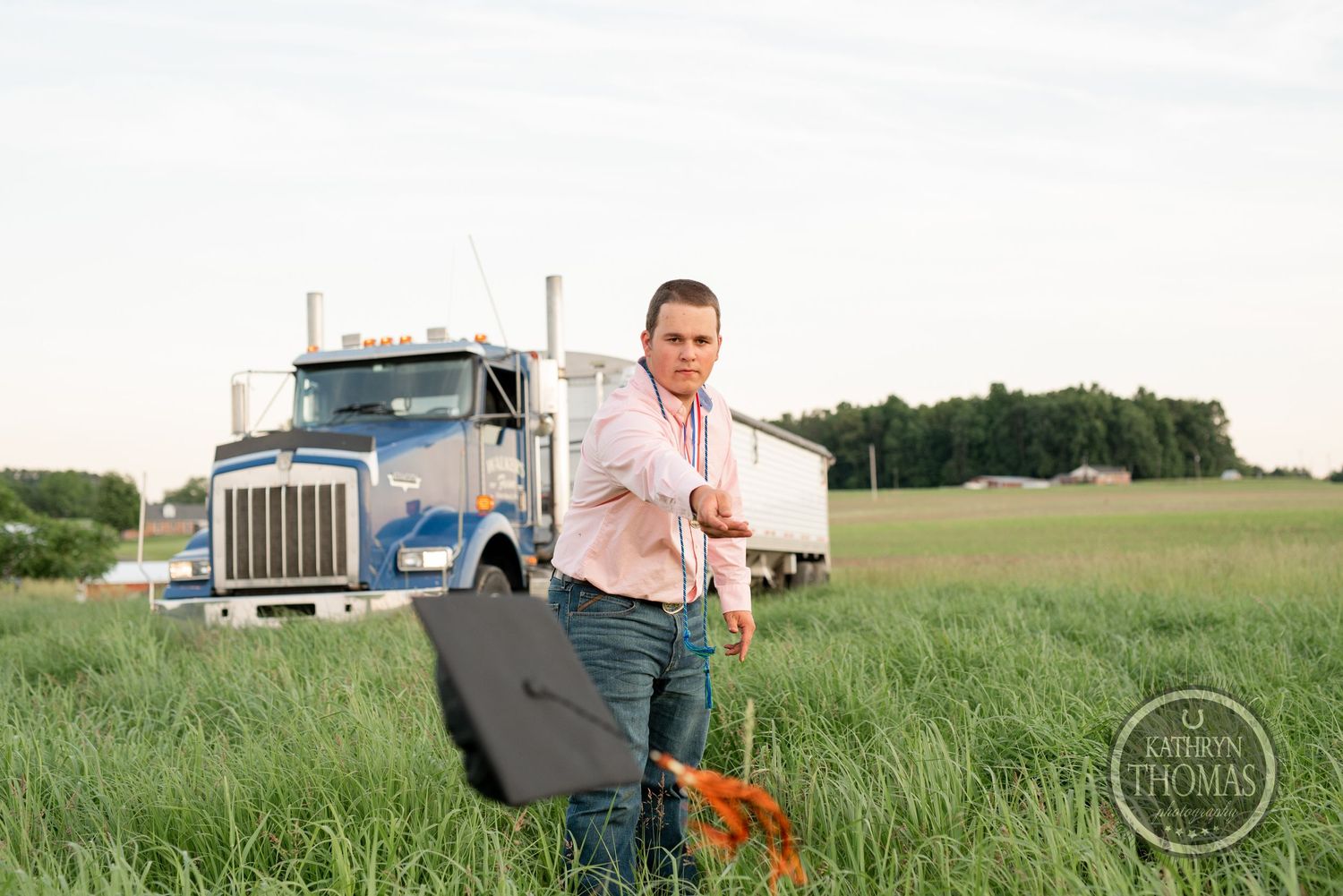 Graduate in pink shirt carrying graduation accessories poses by semi truck in field.