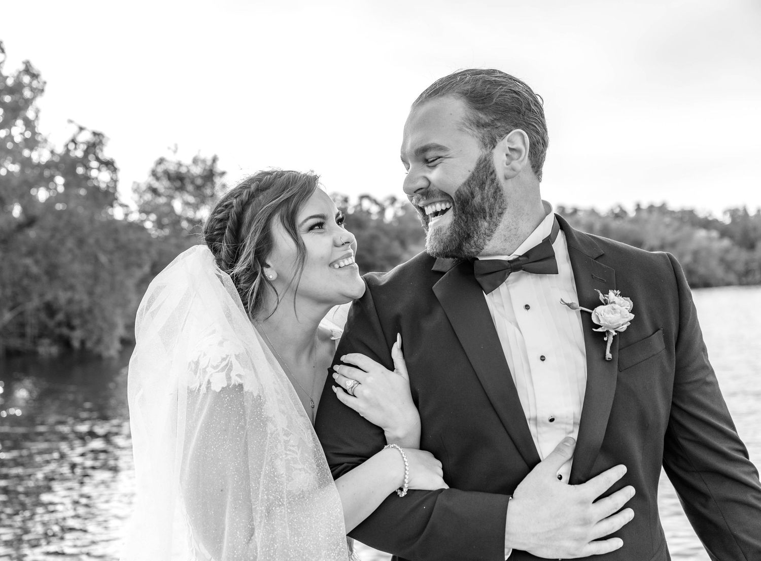 A black and white portrait of a couple in wedding attire laughing together outdoors by a lake.