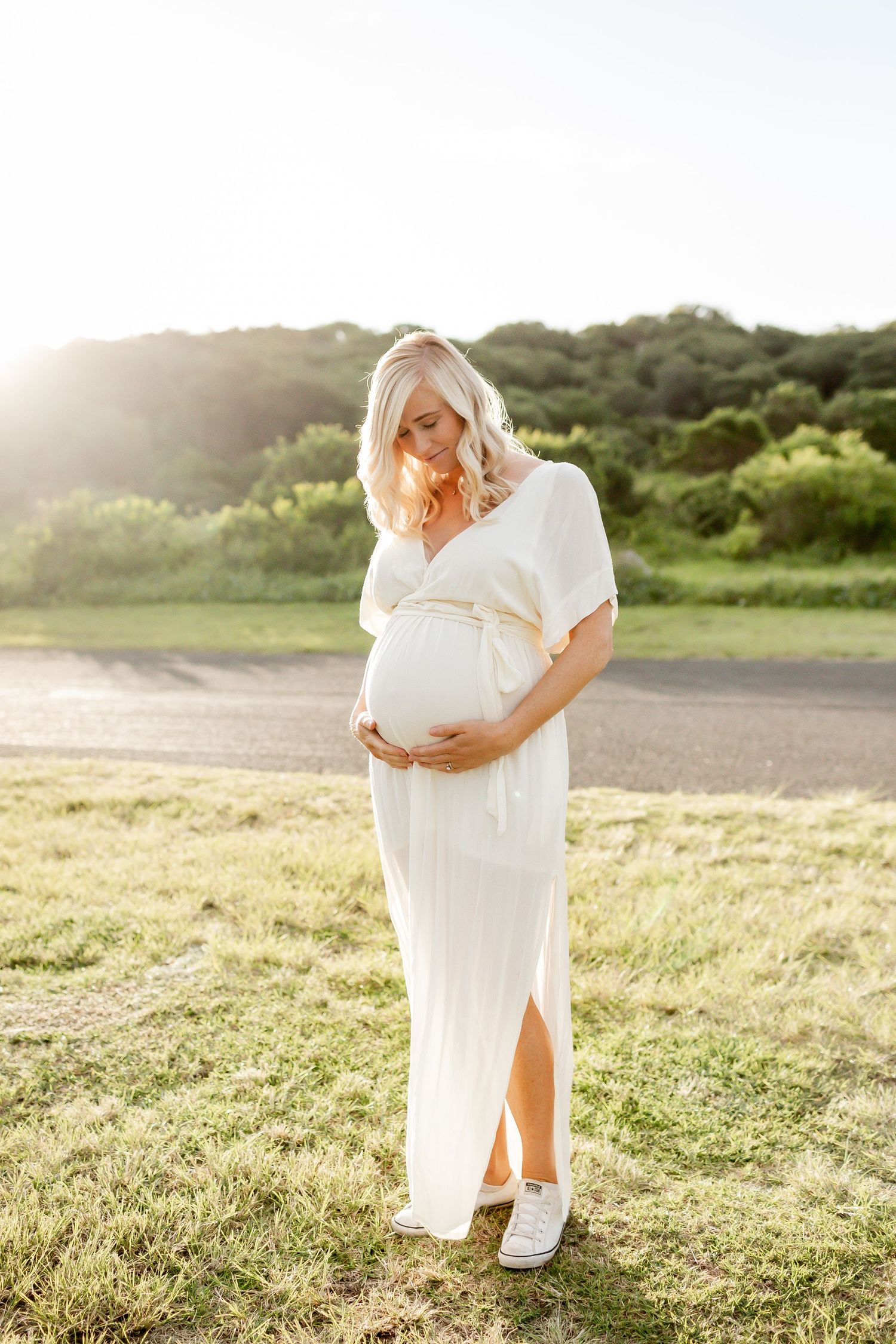 Expectant woman in white dress touching belly during sunset photoshoot.