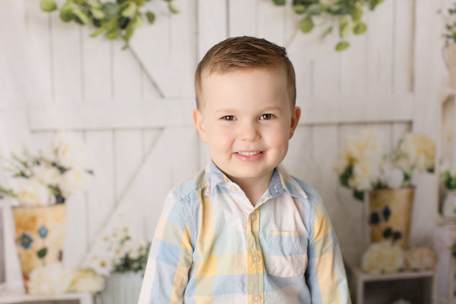 Young child in light blue button-up shirt smiles against white floral backdrop.