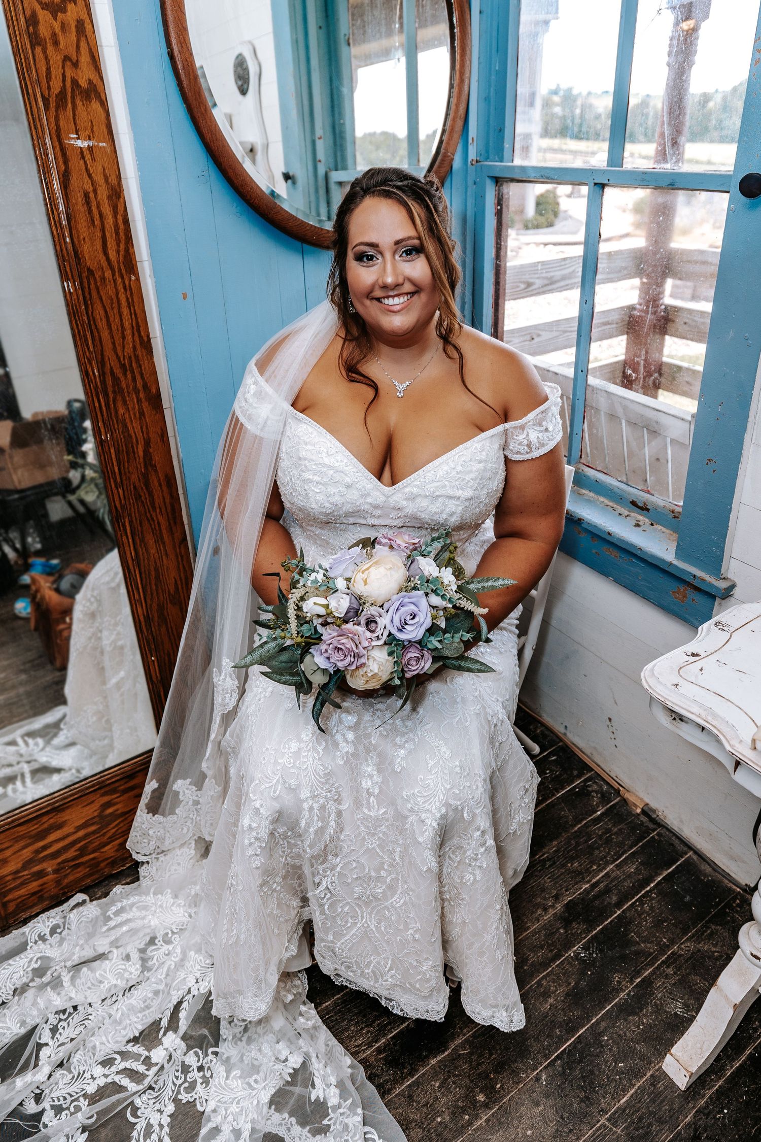 A bride in an off-shoulder lace gown sits with her bouquet in a room with blue walls and vintage details.