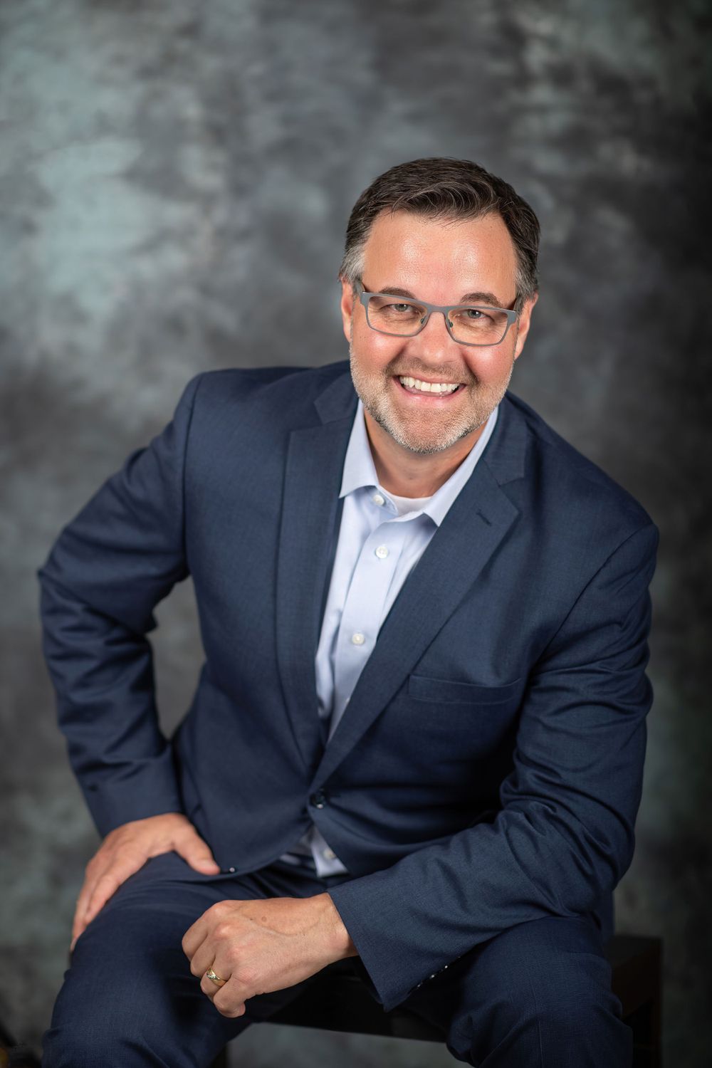 Professional headshot of person in navy suit against dark background.