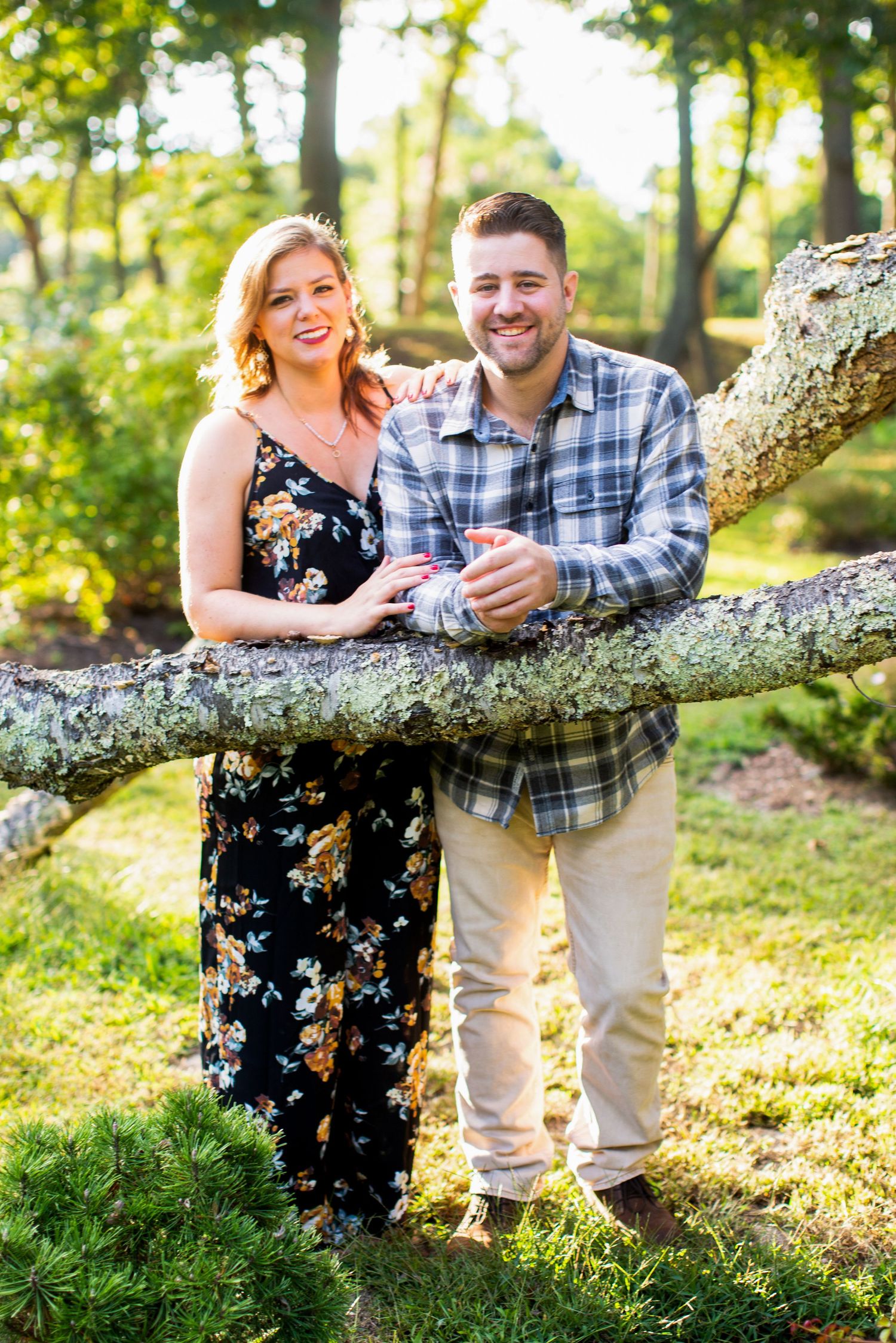 A couple shares a joyful moment while posing on a fallen tree trunk in a sunny outdoor setting.