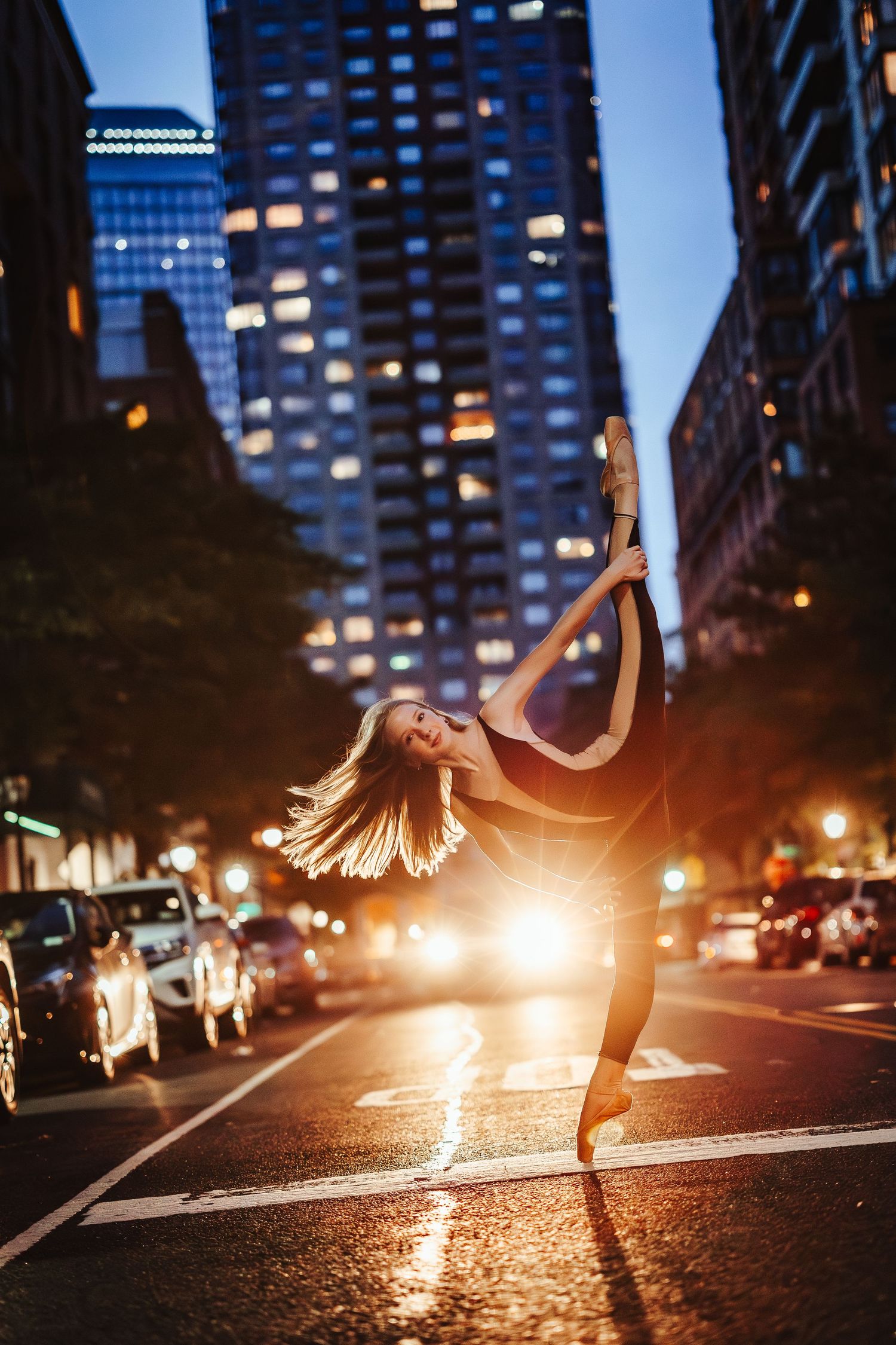 A dancer leaps gracefully on a dark city street illuminated by warm streetlights and towering buildings.