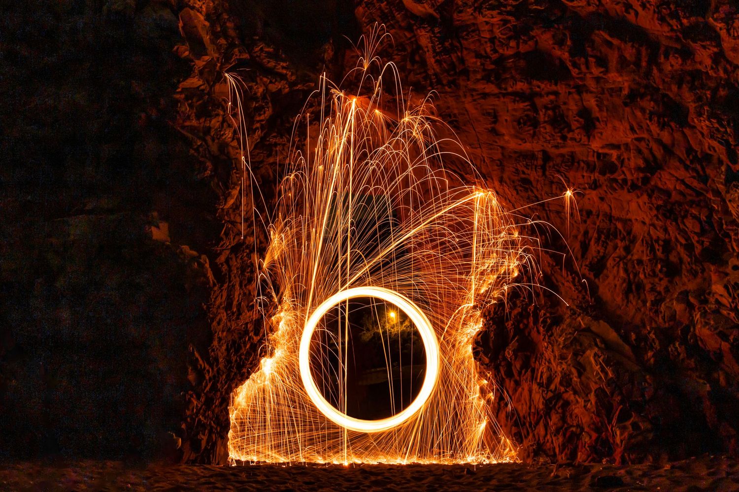 Long-exposure photograph of a glowing ring of fire created by spinning steel wool inside a cave, casting sparks against the rocky walls.