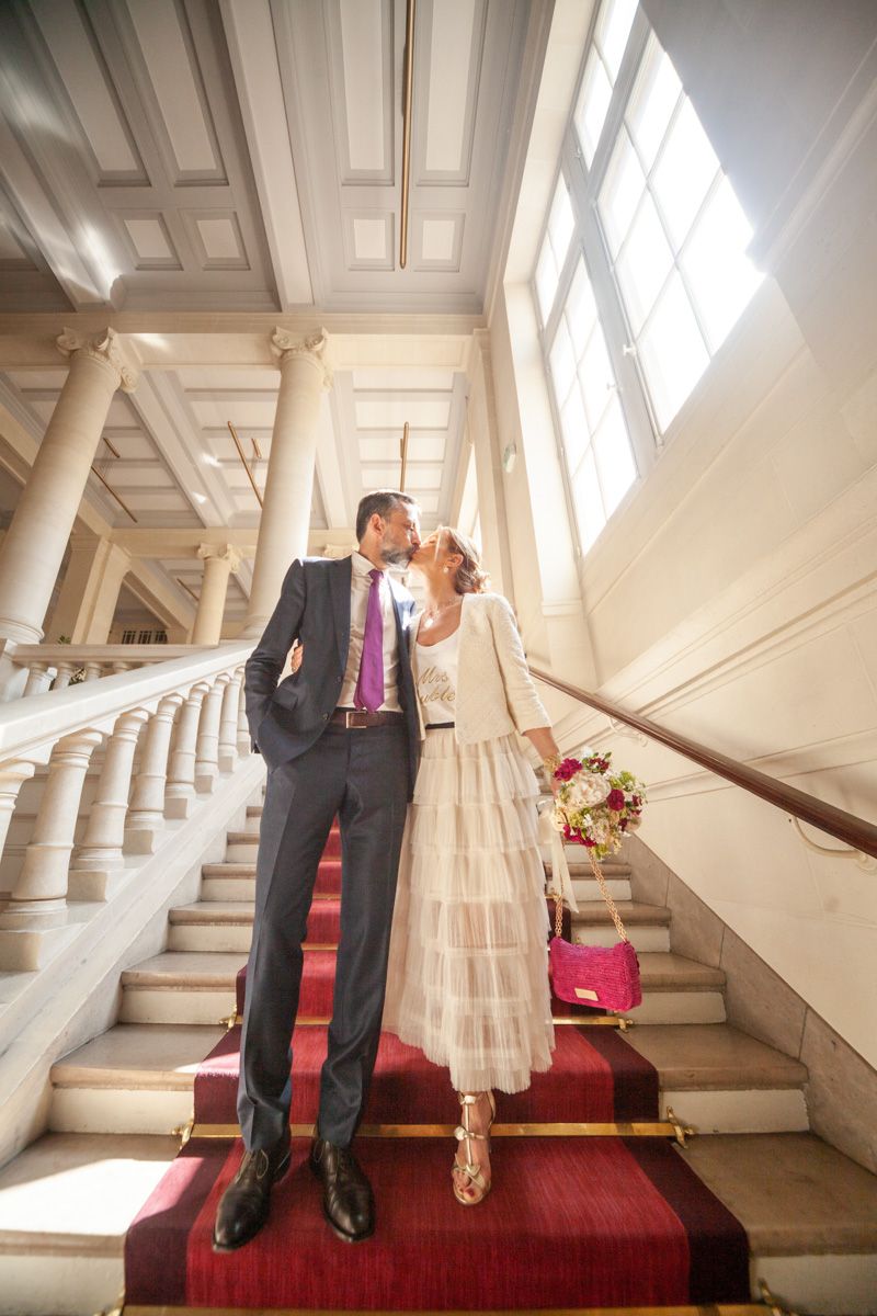 Elegant couple shares romantic moment on grand staircase with sunlight streaming through windows.