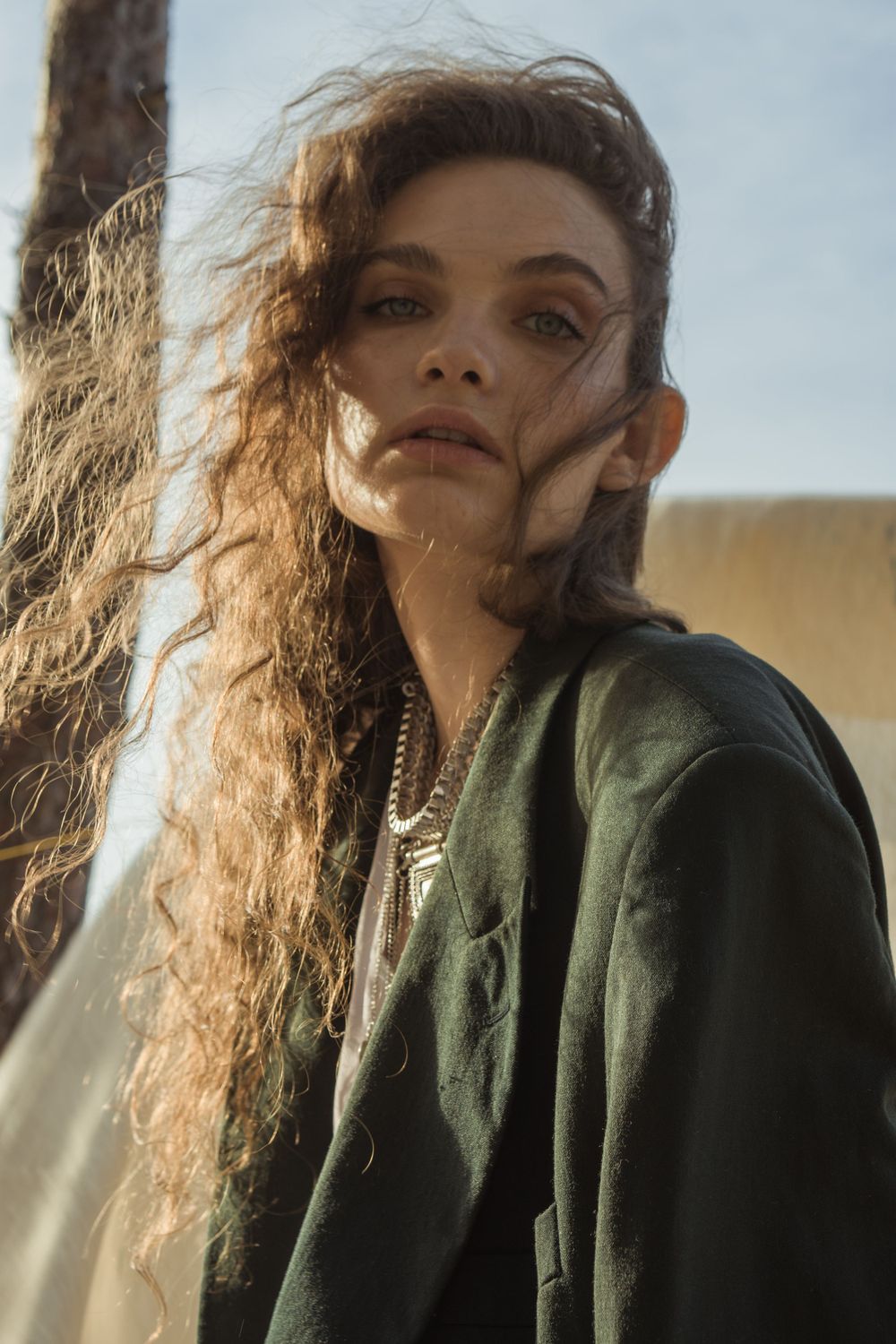 A woman with wavy hair in a dark green blazer, wearing silver necklaces, gazing into the camera in warm sunlight in Soho, LOndon, UK