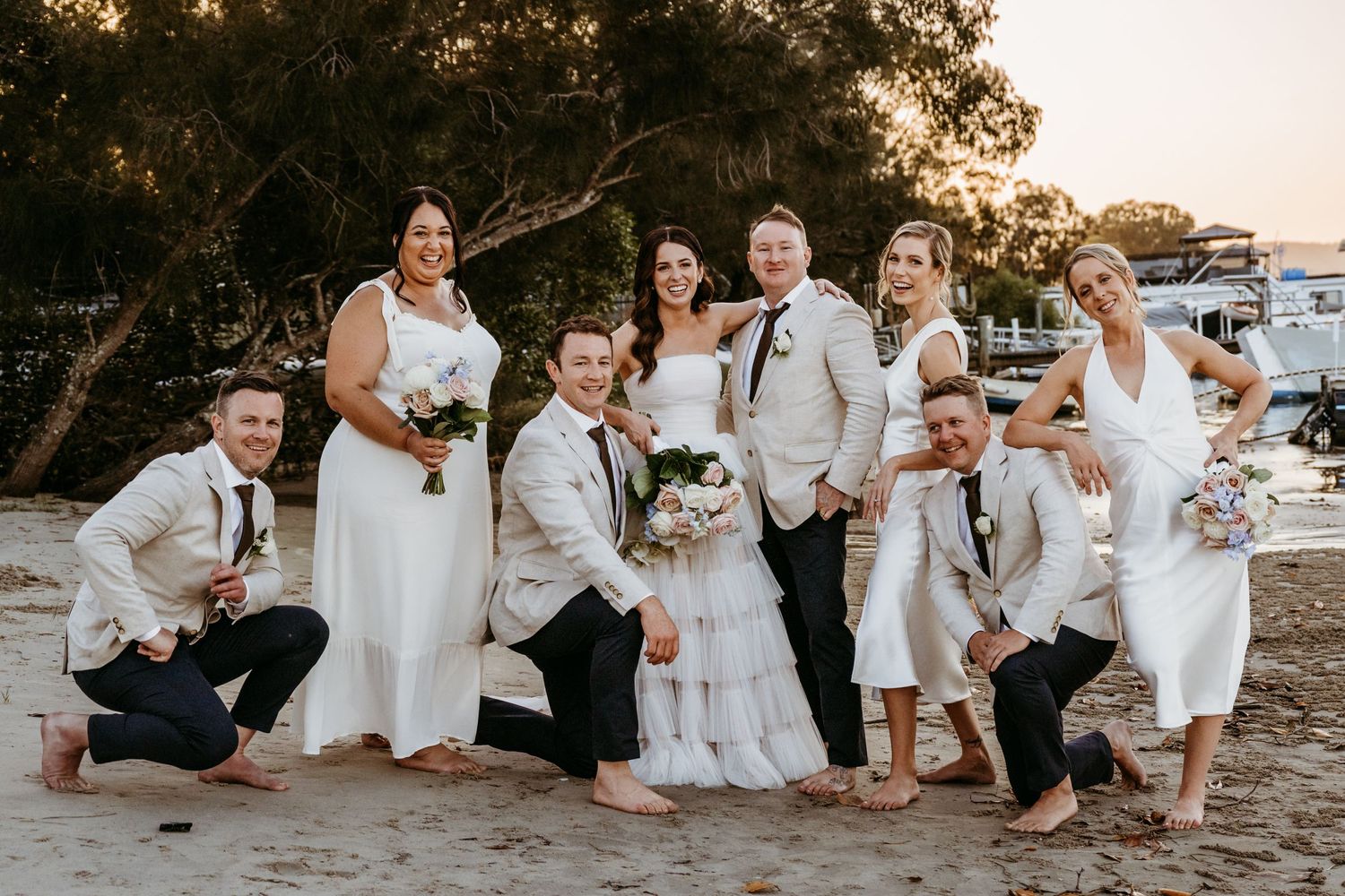 Wedding party poses together on a beach at sunset in coordinated white and cream outfits.