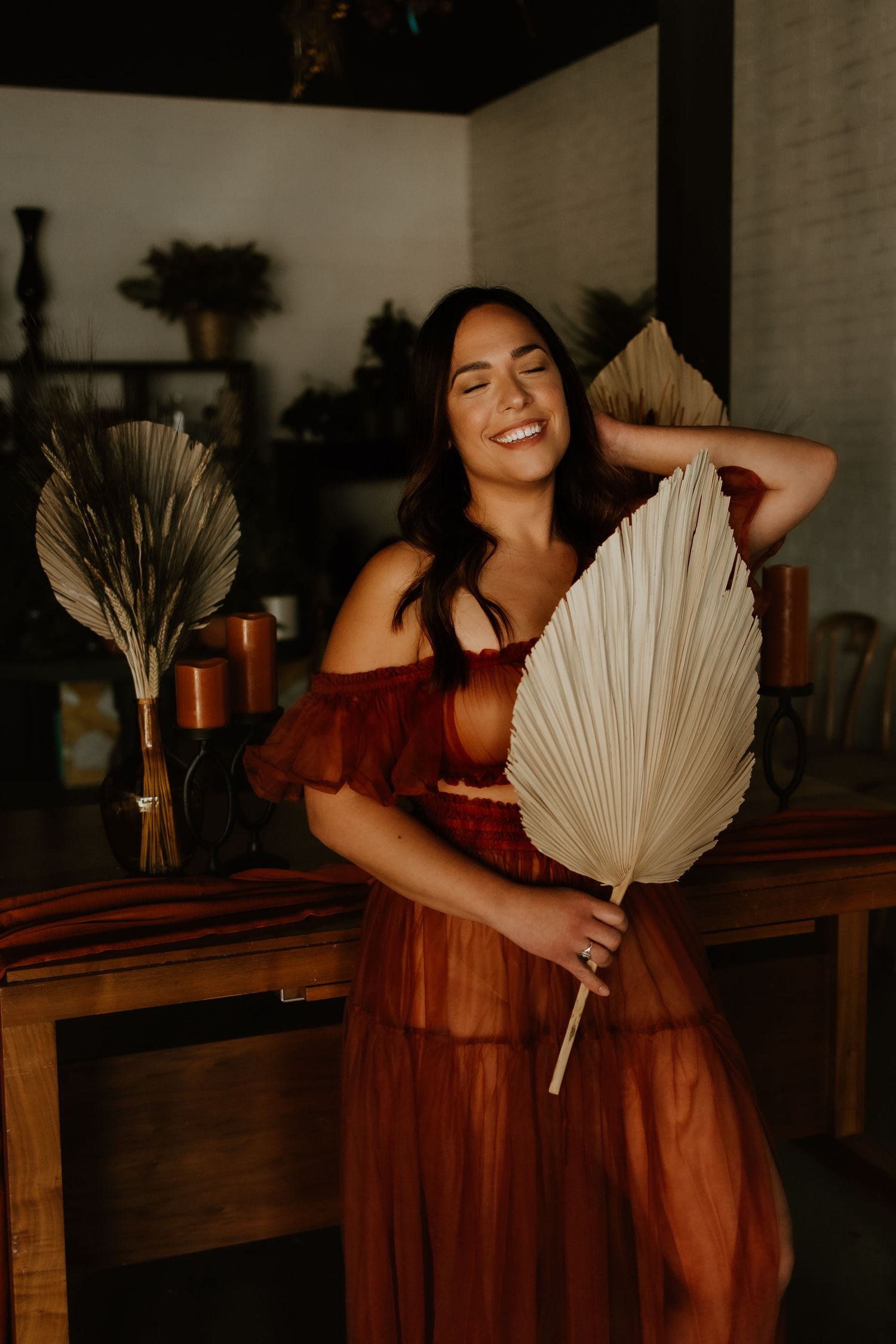 A woman in a burgundy dress poses with dried palm leaves in a boho-styled indoor setting.