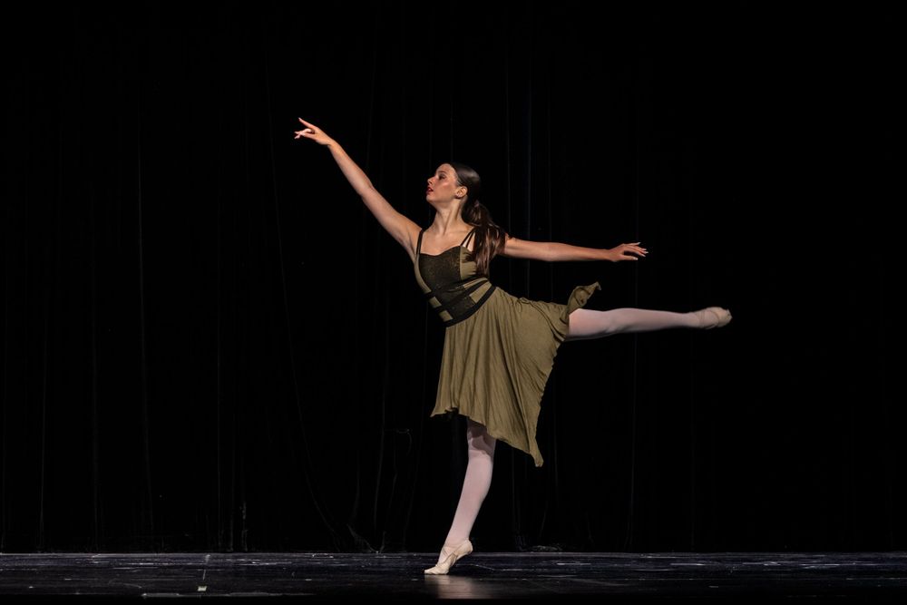 Ballet dancer in olive dress performs graceful arabesque pose on dark stage.