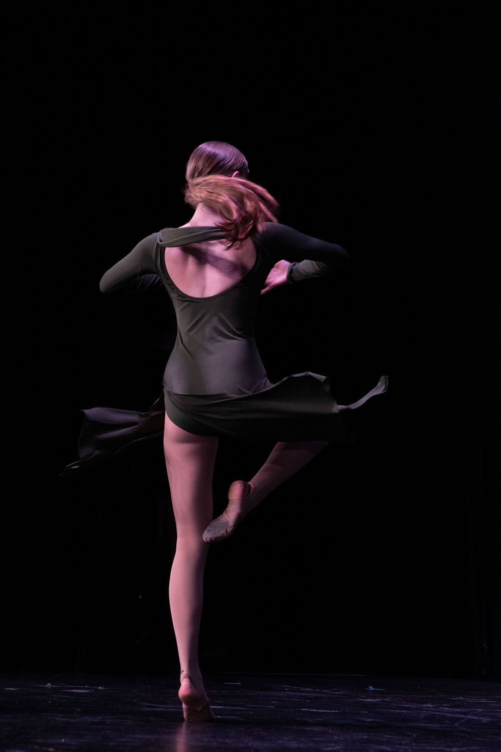 Modern dancer in black costume performs expressive movement on dark stage.