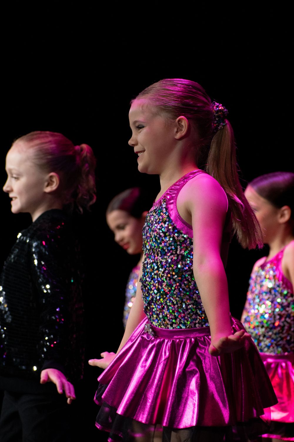 Young dancers in sparkly pink dresses perform in formation on a dark stage.
