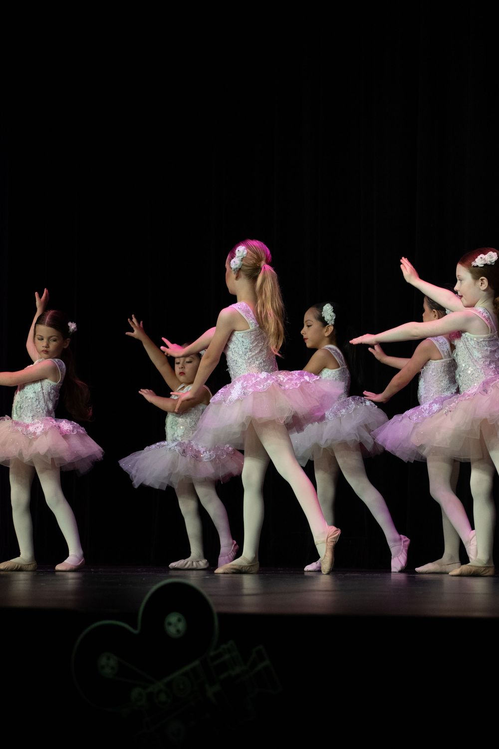 Young ballet dancers in pink tutus perform a choreographed dance routine on a dark stage.