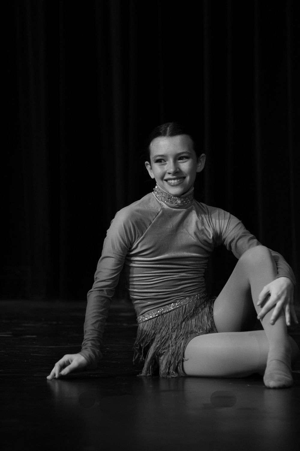 Dancer in fringed costume creates dramatic low pose on dark stage.