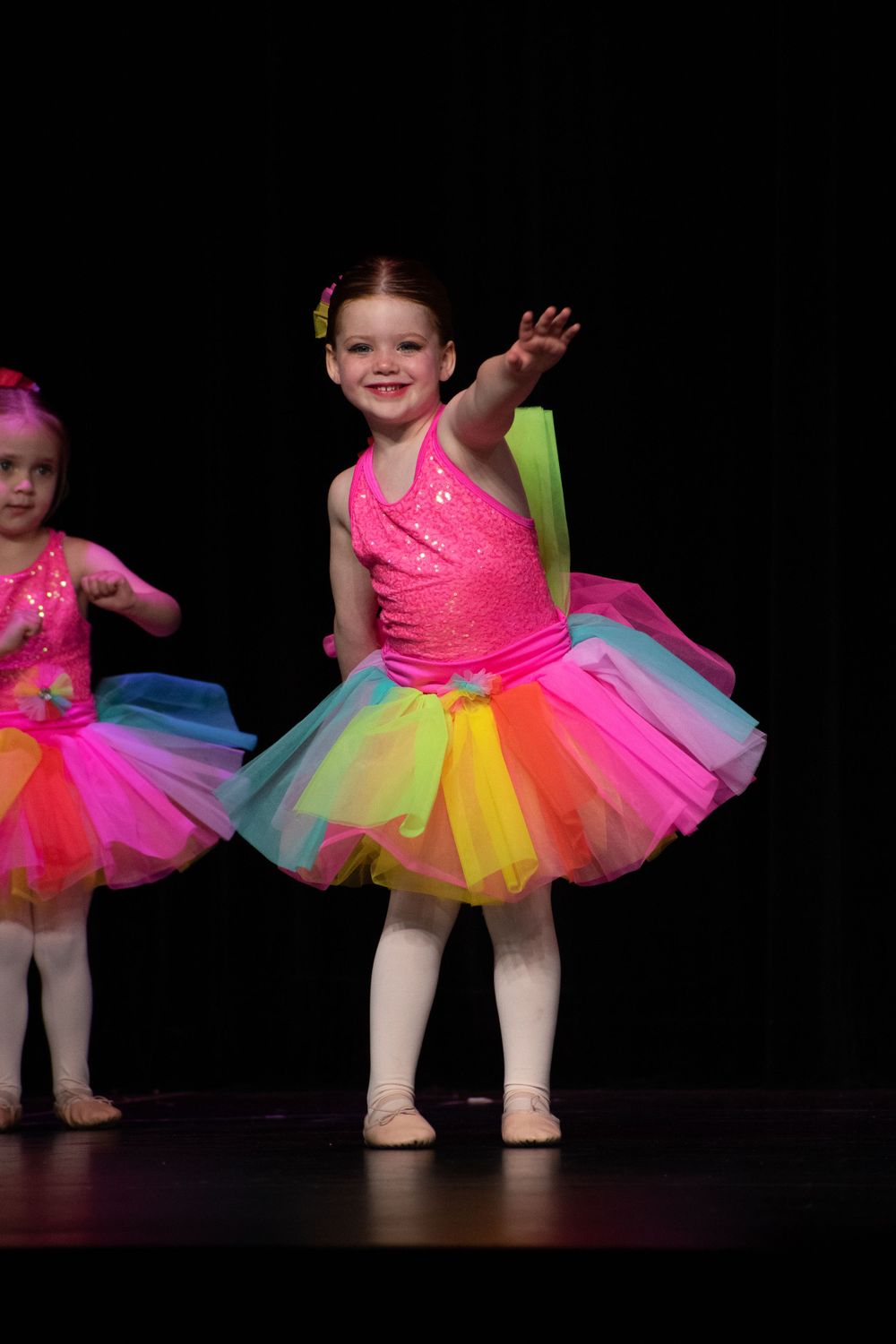 Dancers wearing vibrant rainbow tutus and pink tops perform on a dark stage.