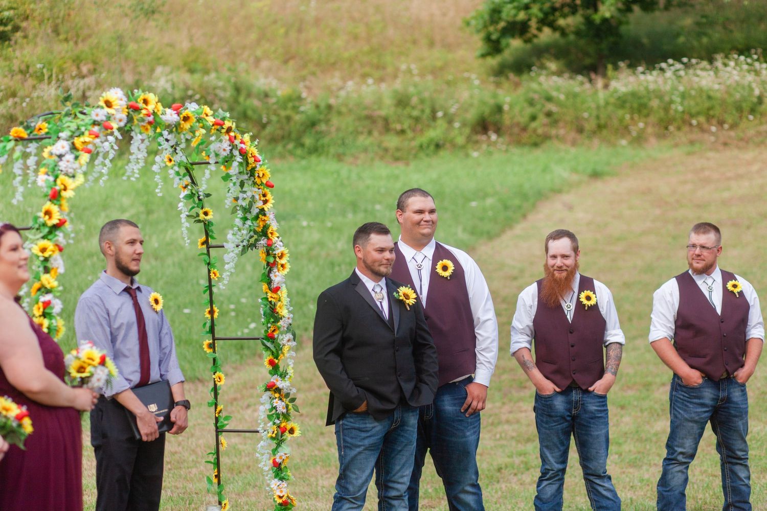 Wedding party stands under a floral arch decorated with sunflowers in an outdoor field setting.