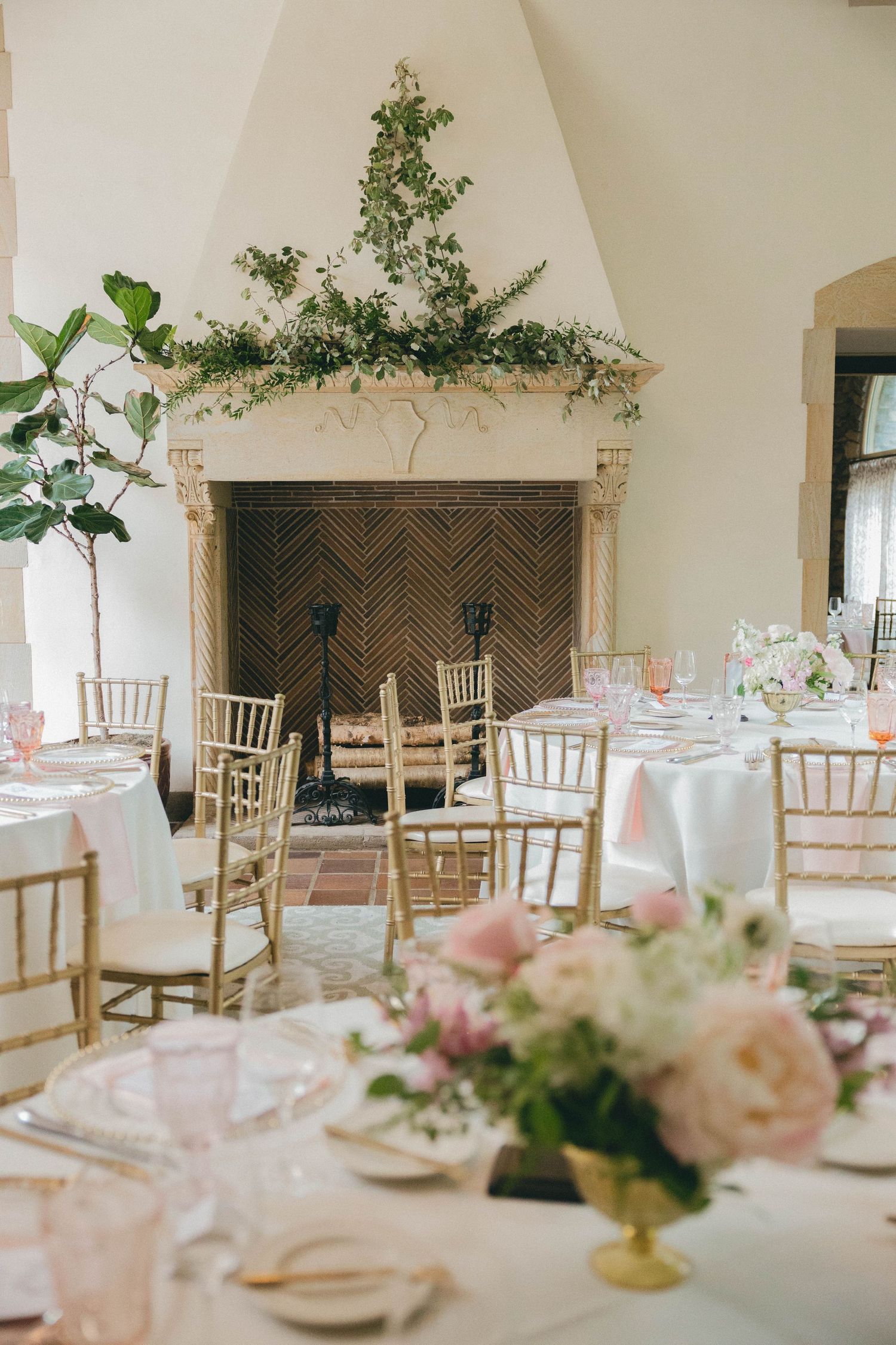 Elegant wedding reception venue with gold chairs, floral centerpieces, and greenery over a fireplace.