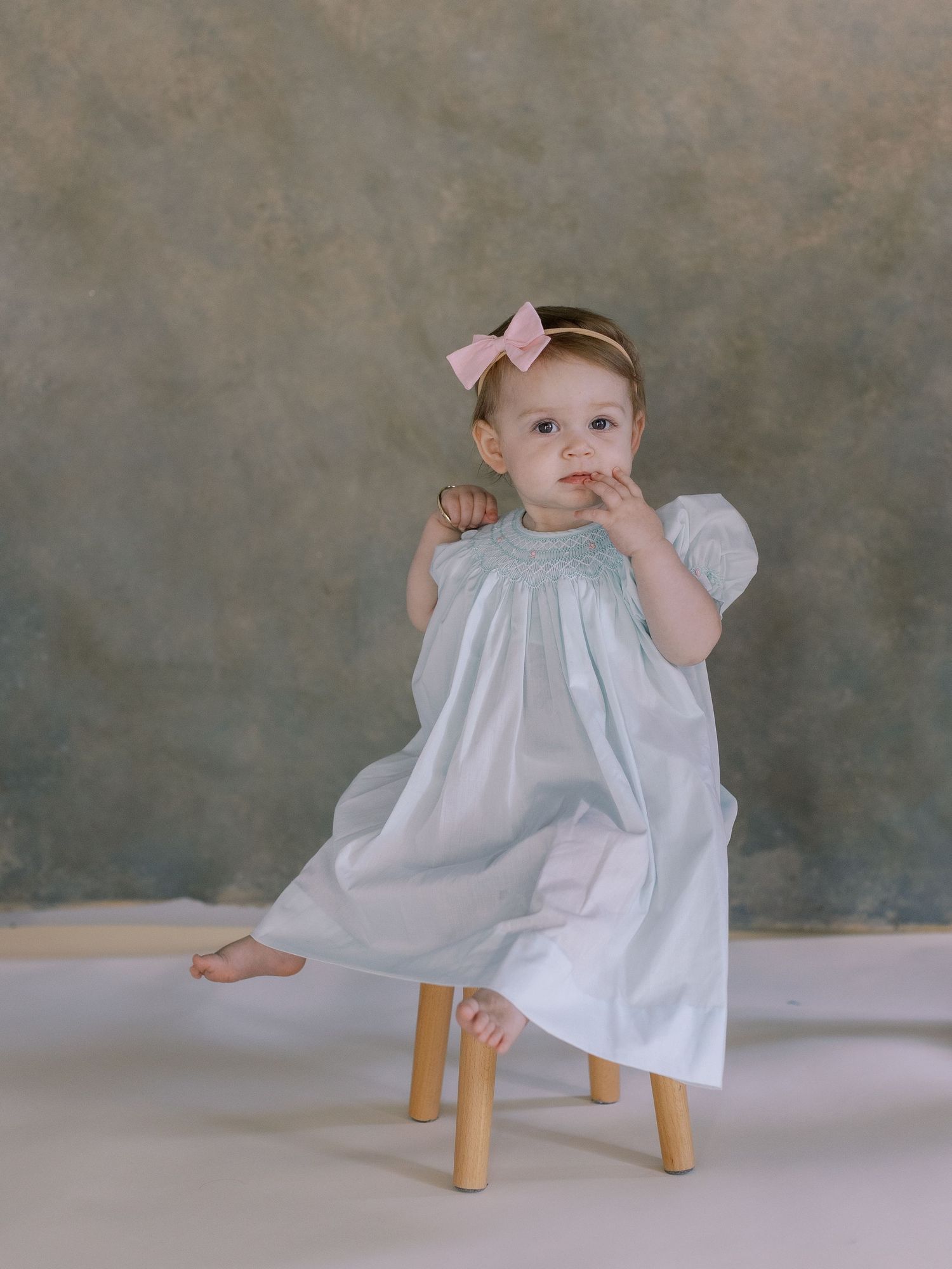Young child in white dress and pink bow sitting on wooden stool against textured backdrop.