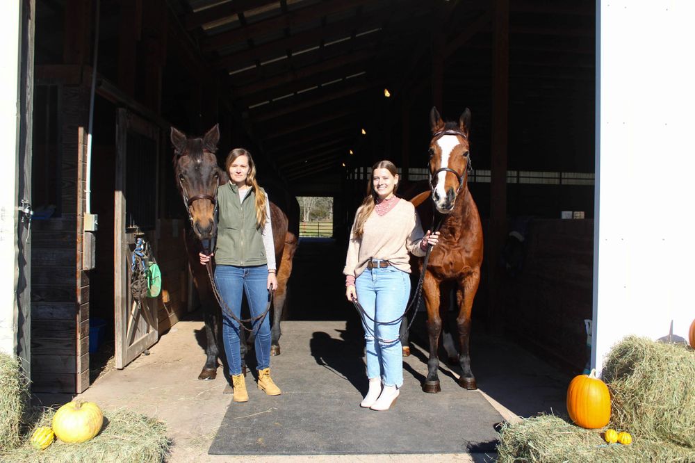 Two people walk with horses inside a barn stable with pumpkin decorations.