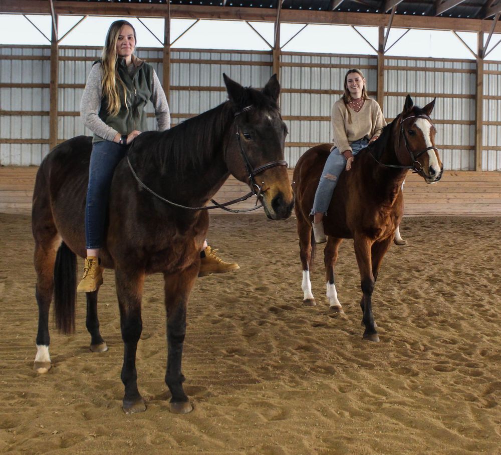 Two horses and their riders standing in a sandy indoor arena.