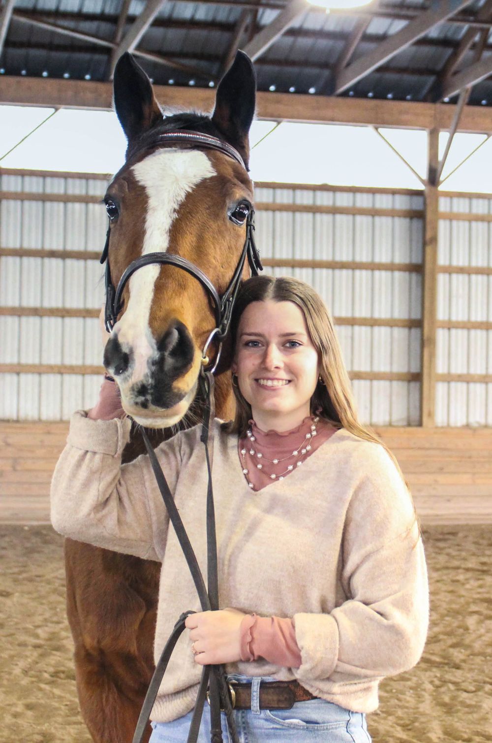 A bay horse with white blaze poses with its rider in an indoor riding arena.
