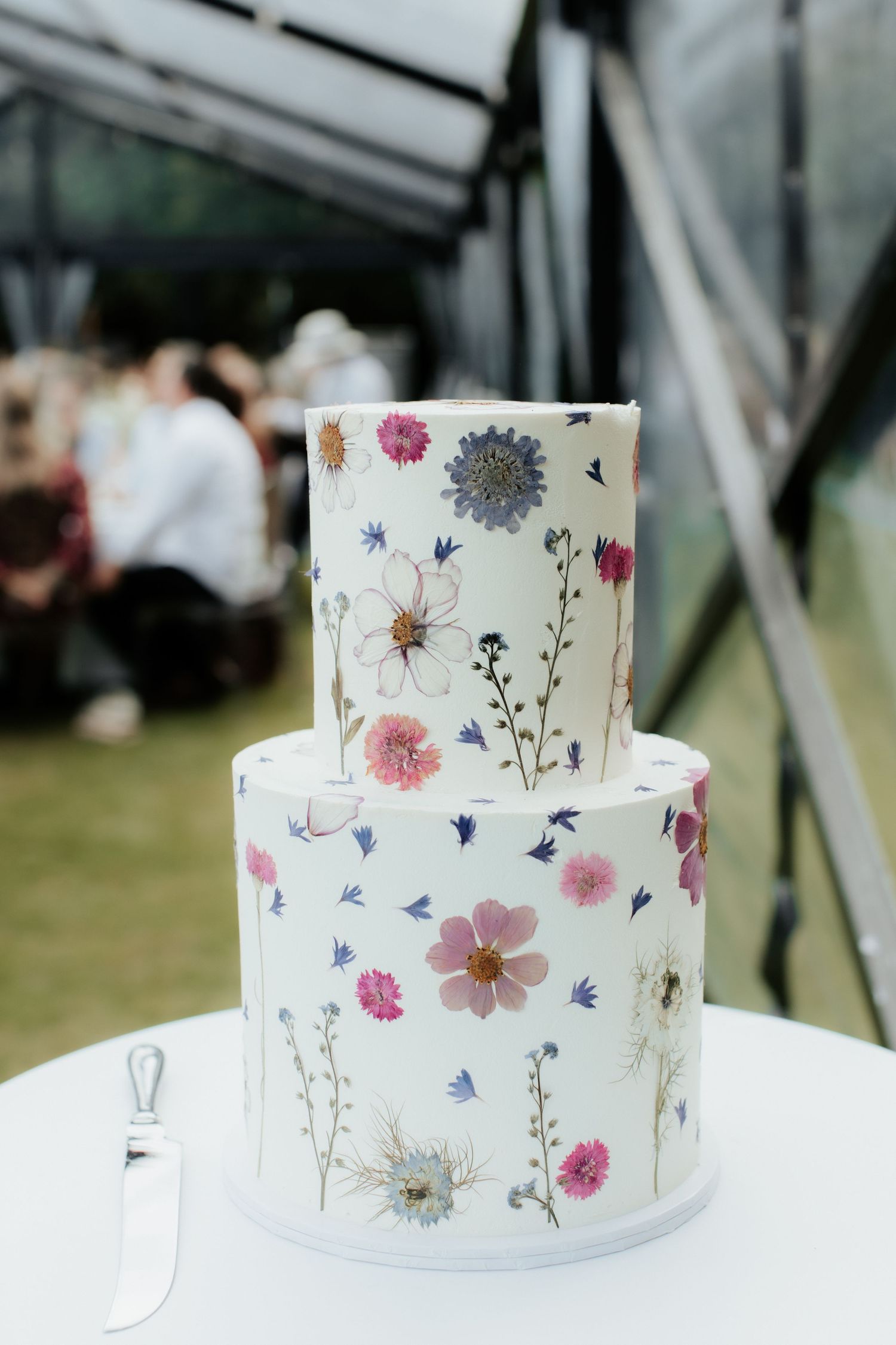 Elegant two-tier wedding cake decorated with pressed wildflowers in pink and blue on white fondant.