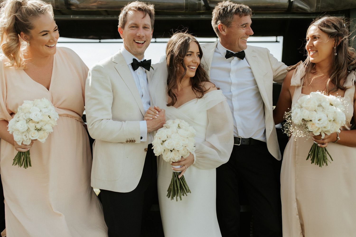 Wedding party members in white and blush pink attire smile together in an informal group photo.