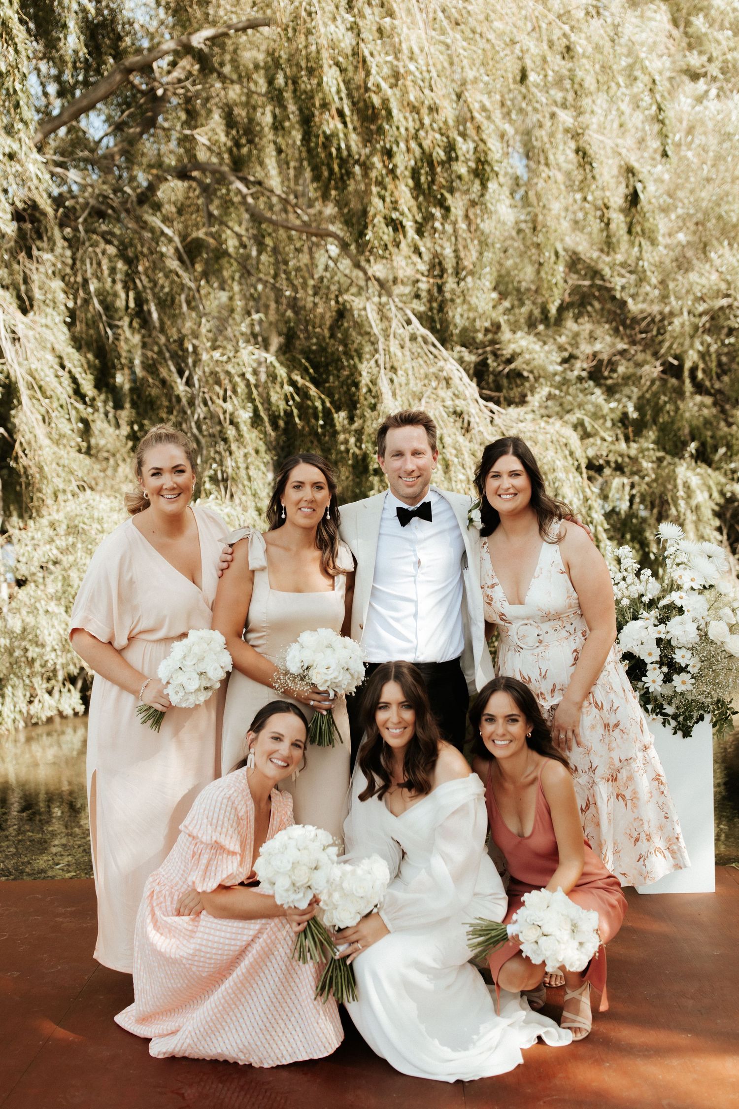 Wedding party poses together outdoors under a weeping willow tree with white floral bouquets.