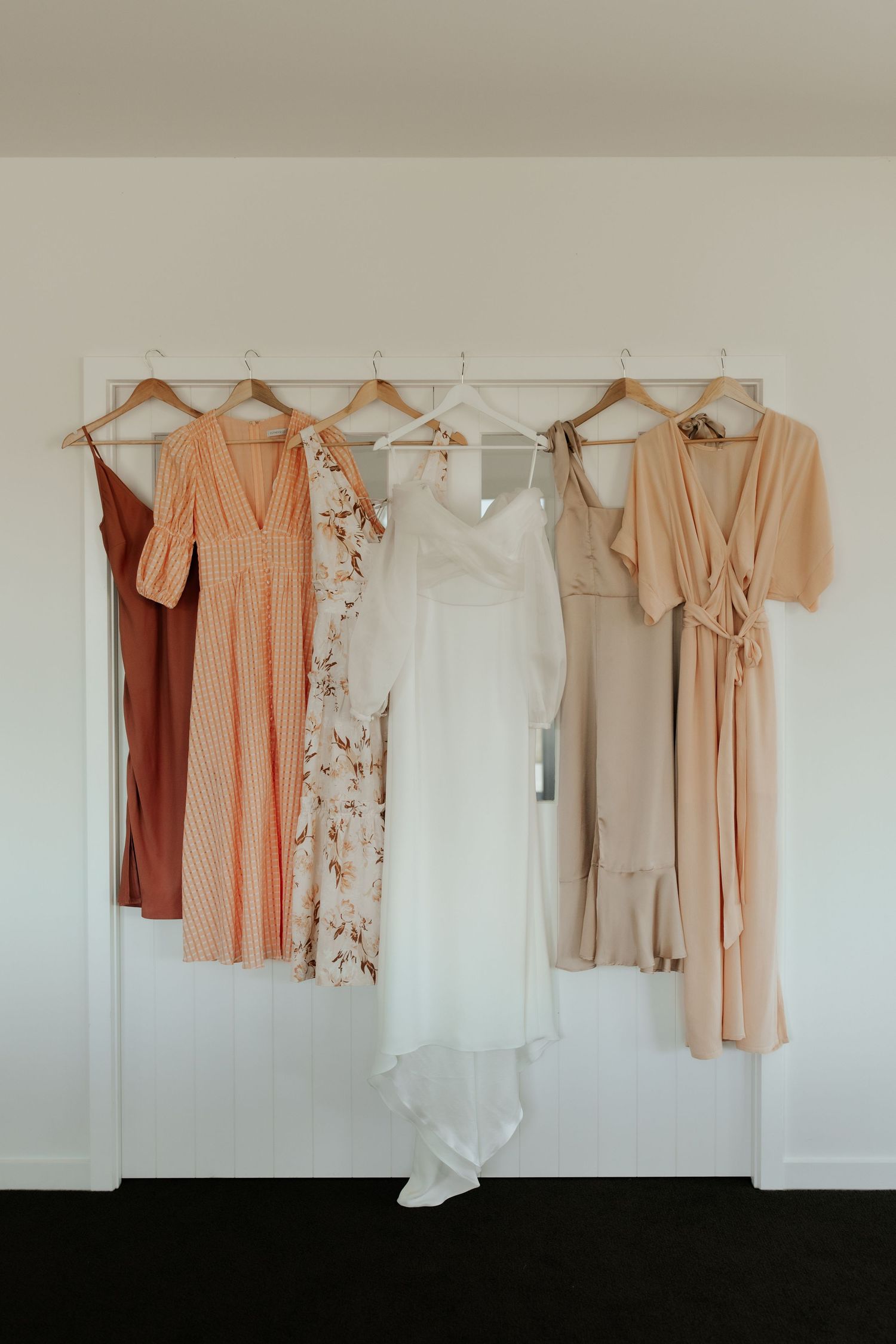 Wedding dress and bridesmaid dresses in peach and neutral tones hanging on wooden hangers.