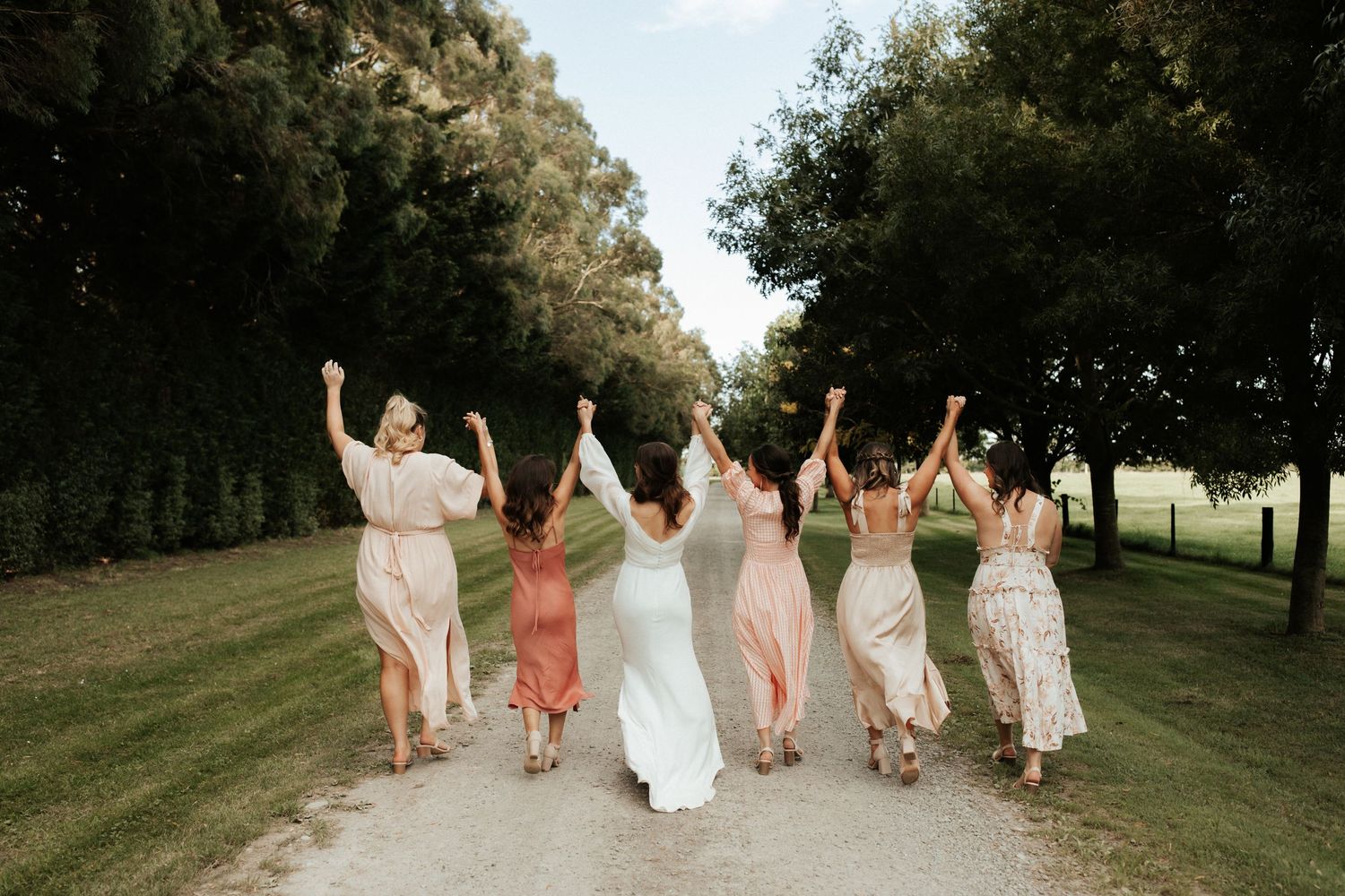 Bridesmaids in blush dresses celebrate joyfully with raised arms on a tree-lined path.