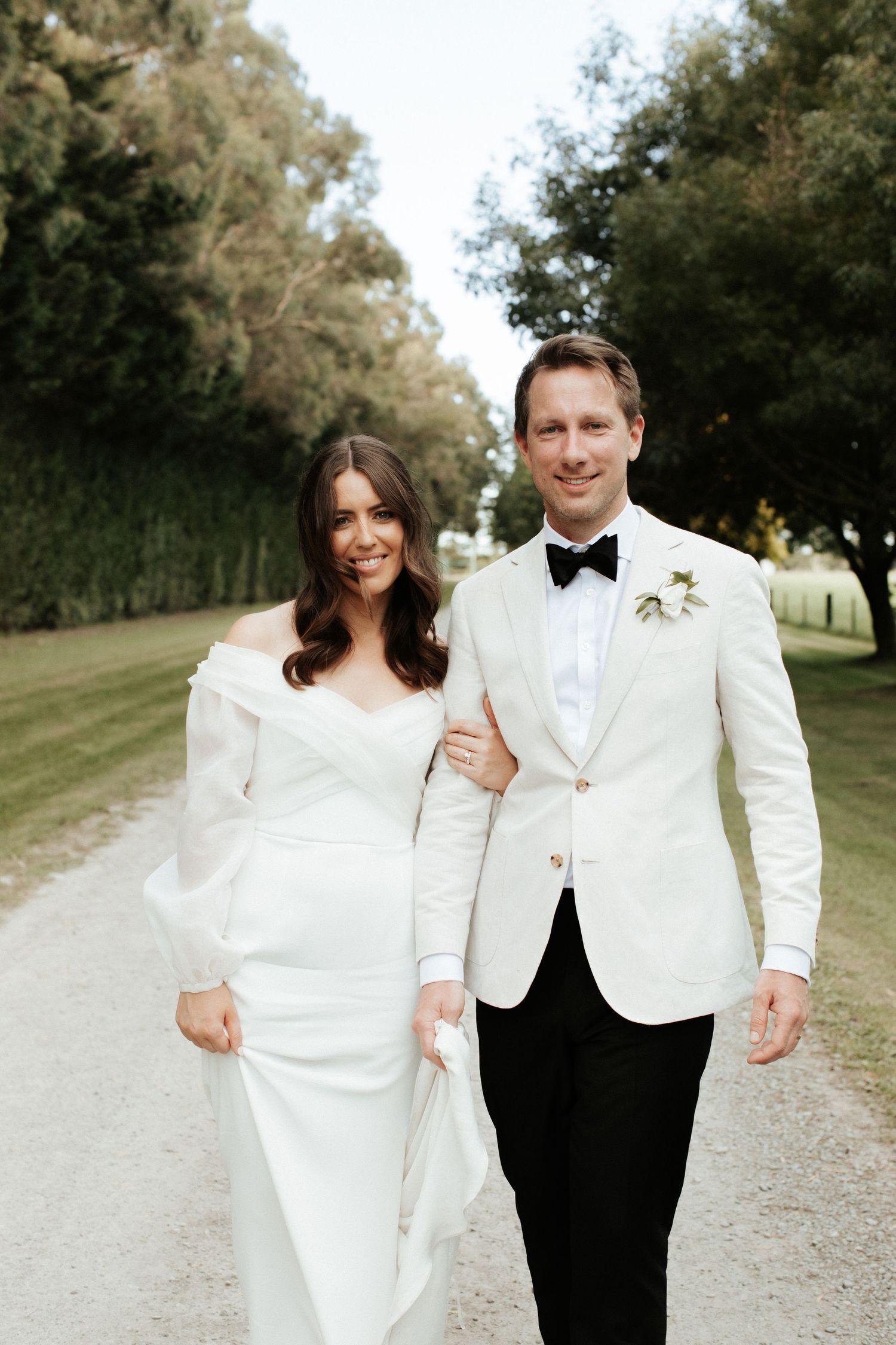 Couple poses together on a tree-lined path, with groom in white jacket and bride in elegant gown.