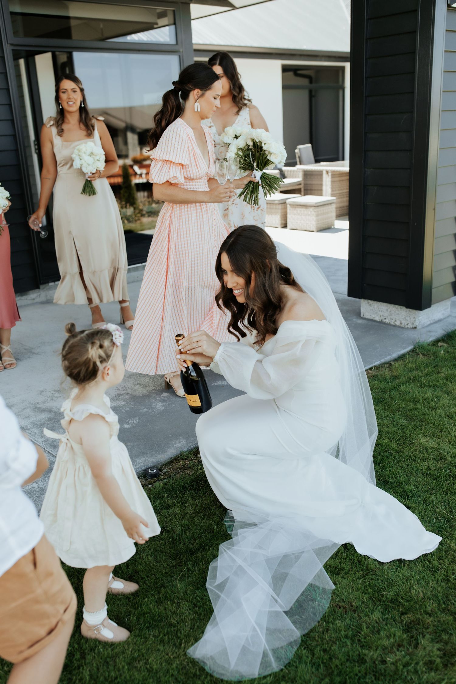A bride in a white dress kneels on grass while opening champagne during an outdoor wedding celebration.