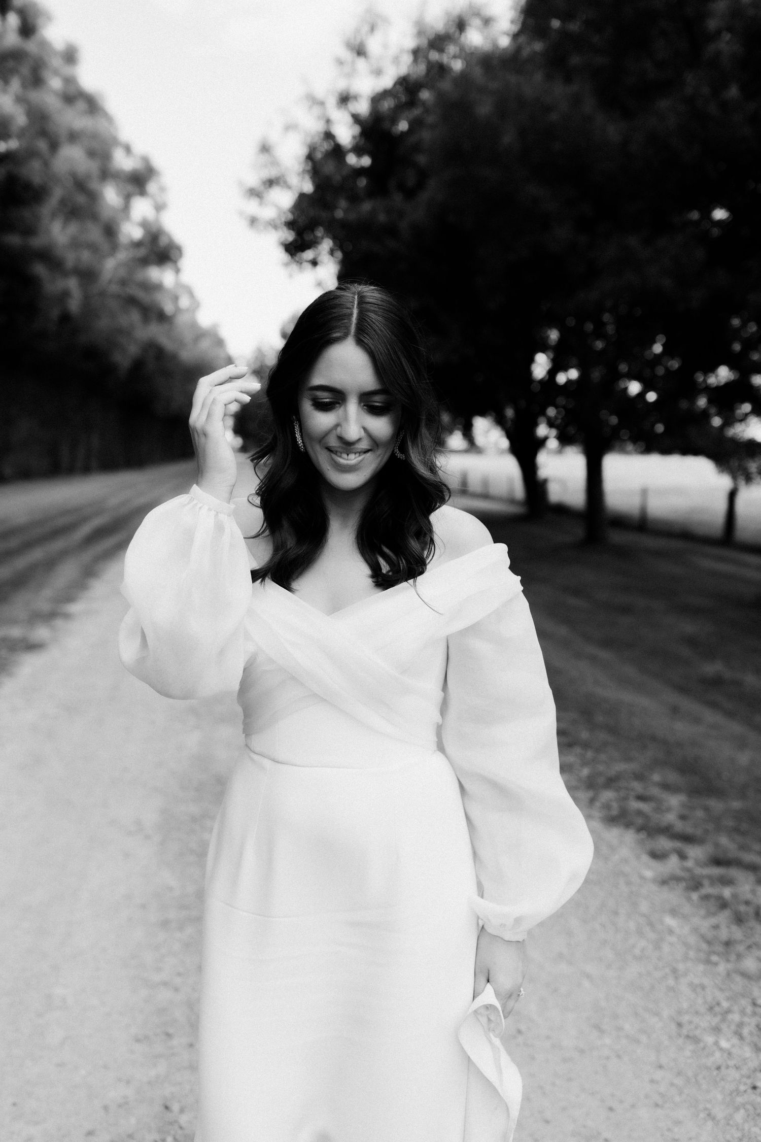 Elegant bride in off-shoulder white gown poses on a tree-lined path in black and white.