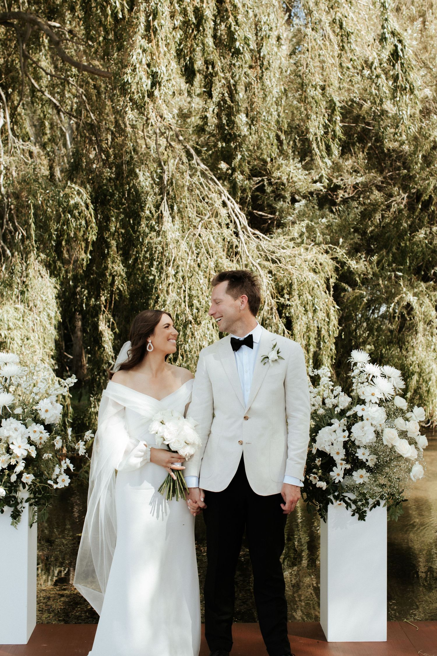 Newlyweds share a joyful moment under a willow tree with elegant white flower decorations.