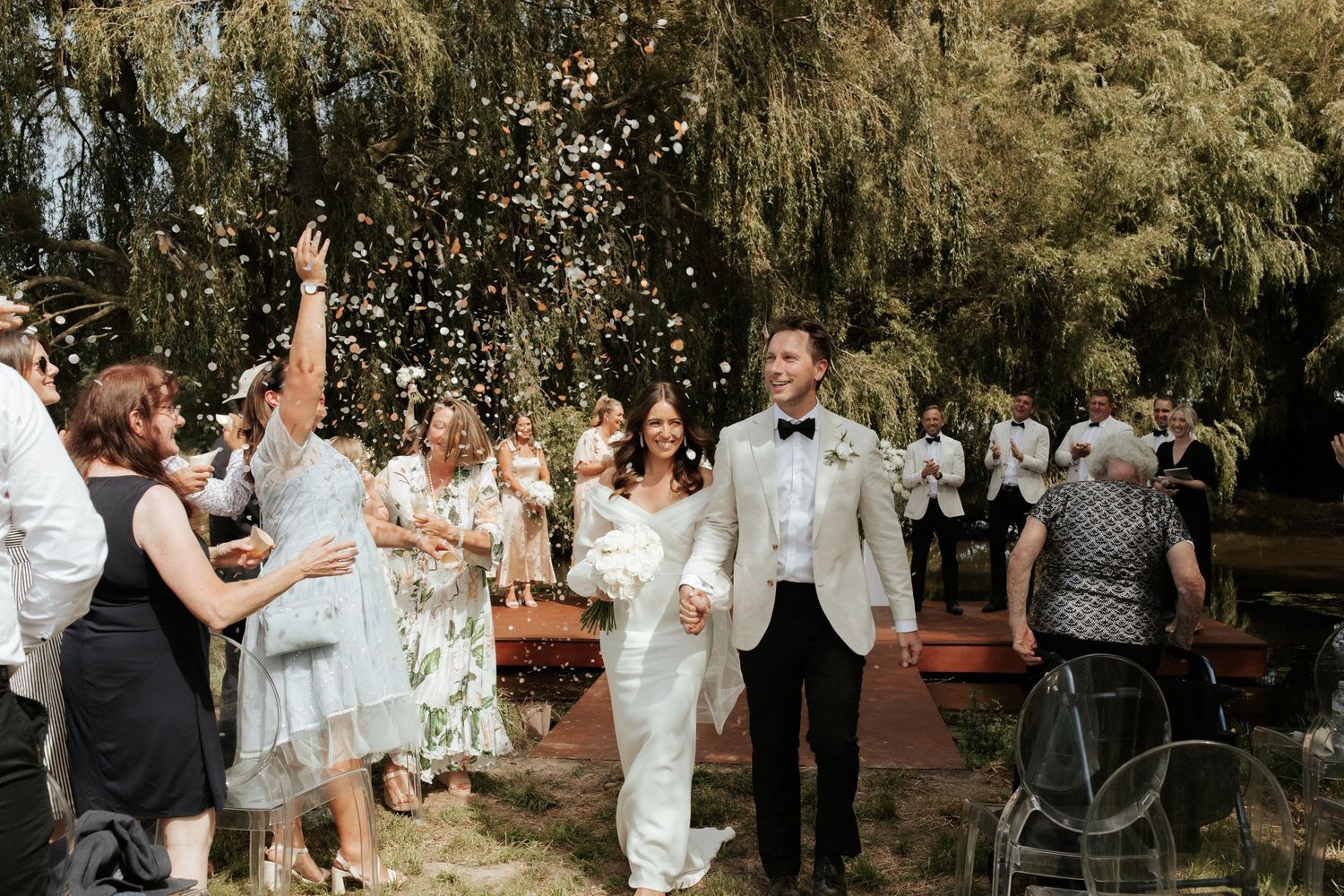 Joyful wedding recessional with guests throwing confetti under willow trees.