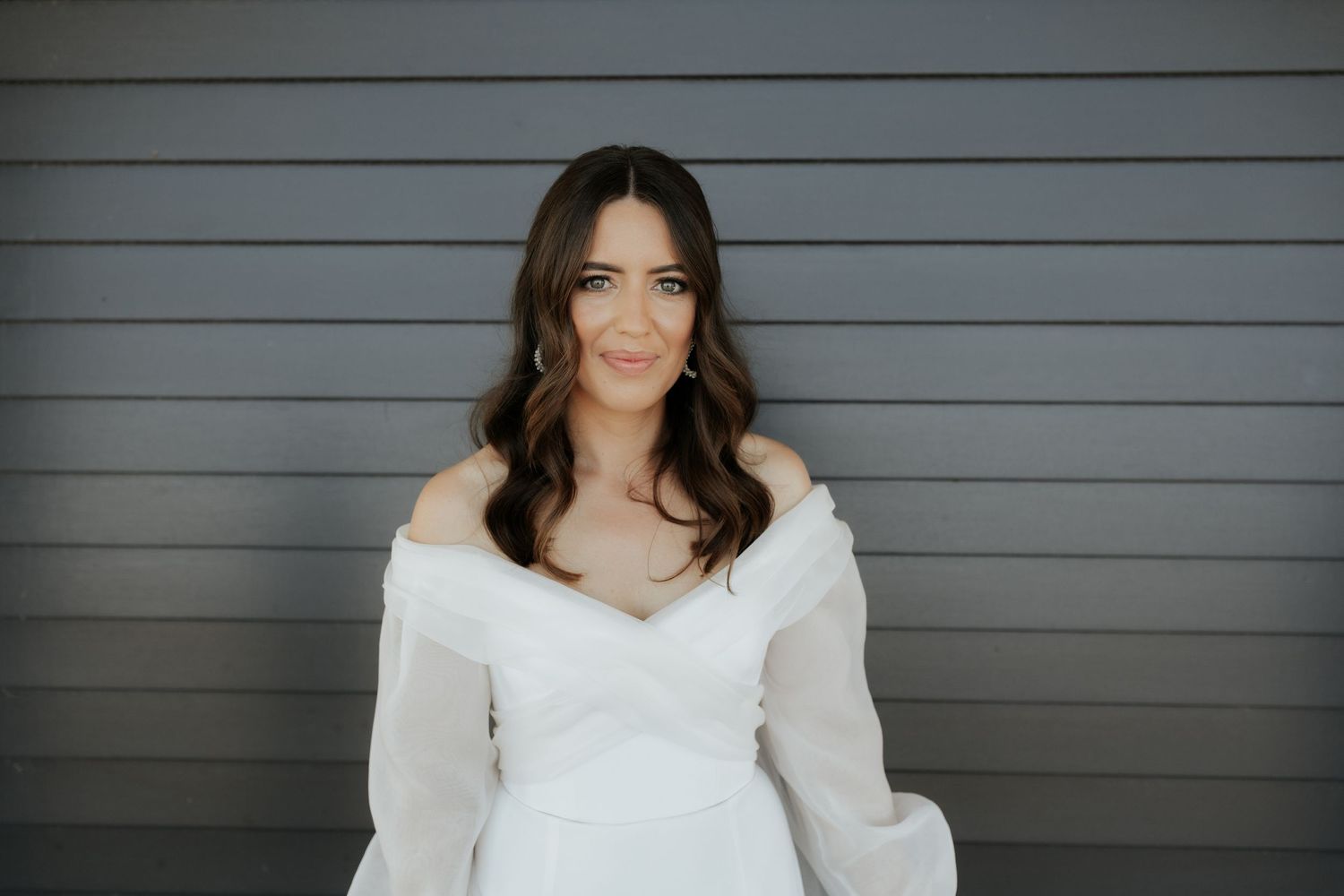 Close-up portrait against gray wall of bride in off-shoulder wedding dress.