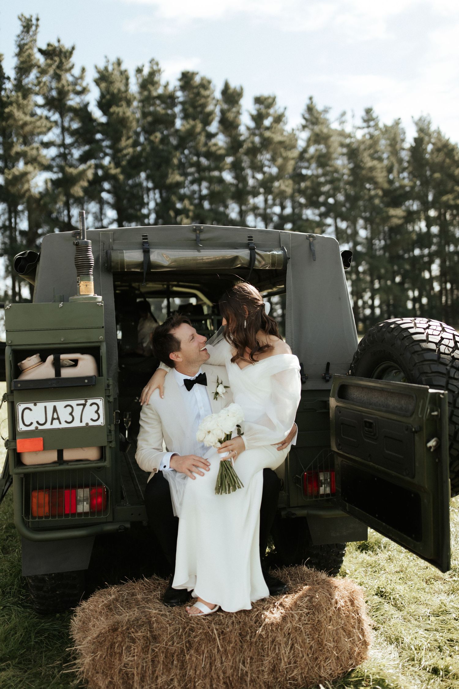 Newlyweds pose romantically on a hay bale in front of a military vehicle.