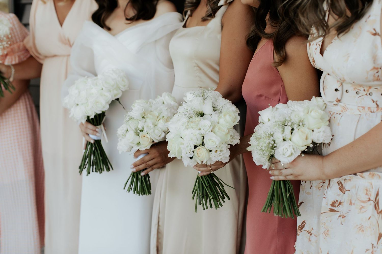 A row of elegant white peony bouquets held against pastel bridesmaid dresses at a wedding.