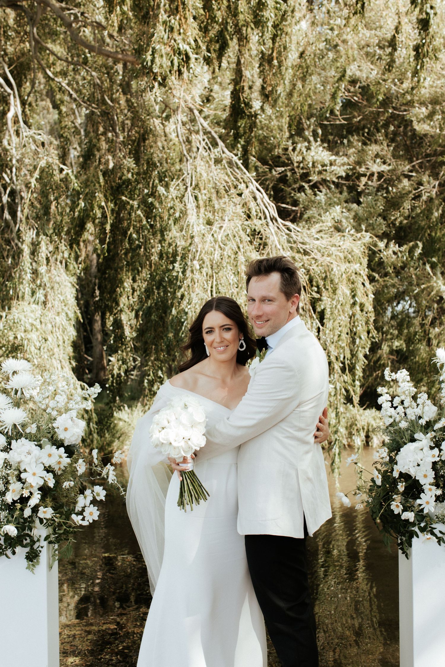 A couple poses under a weeping willow tree with white floral decorations at their outdoor wedding.