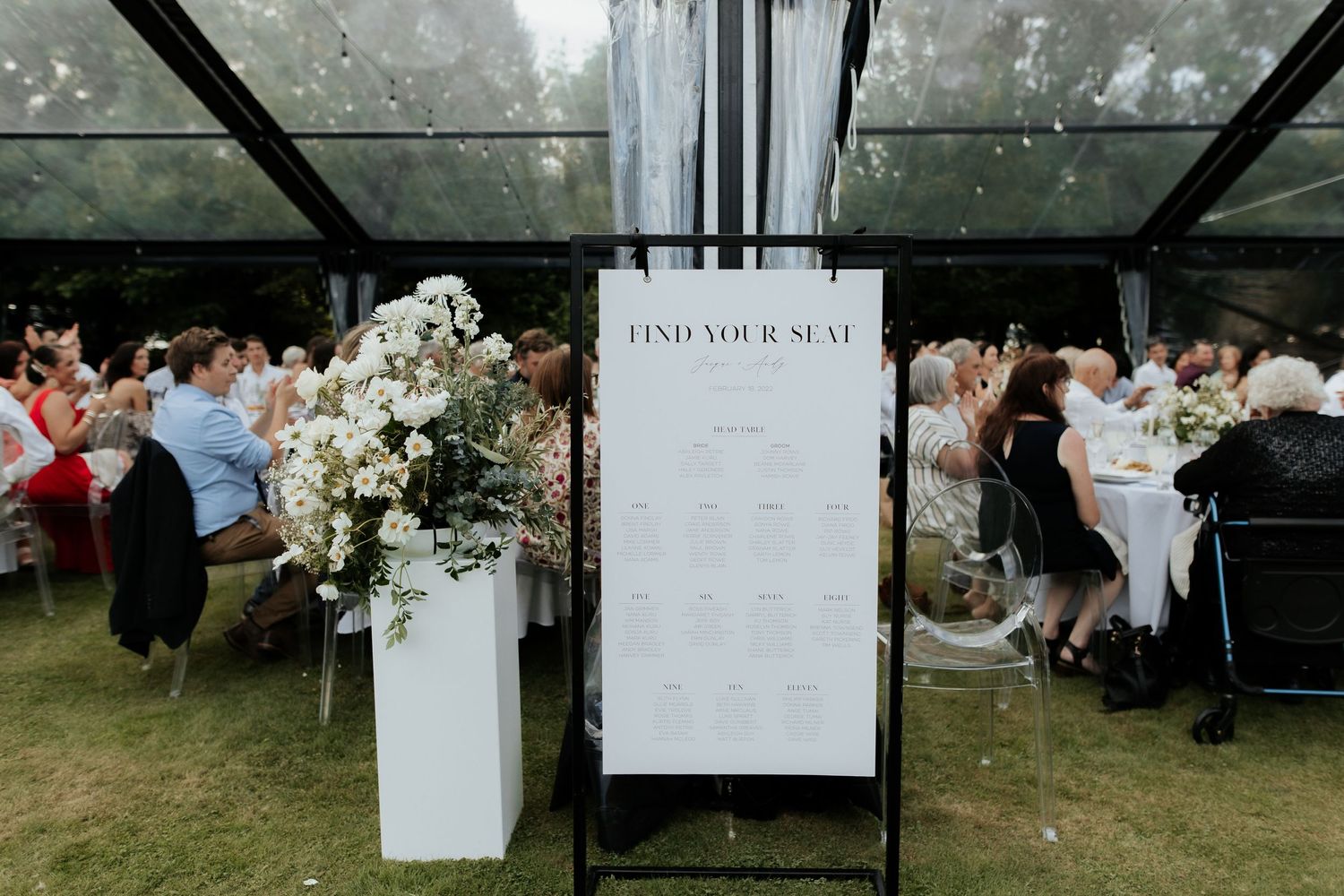 Wedding seating chart displayed on stand with floral arrangement at reception entrance.