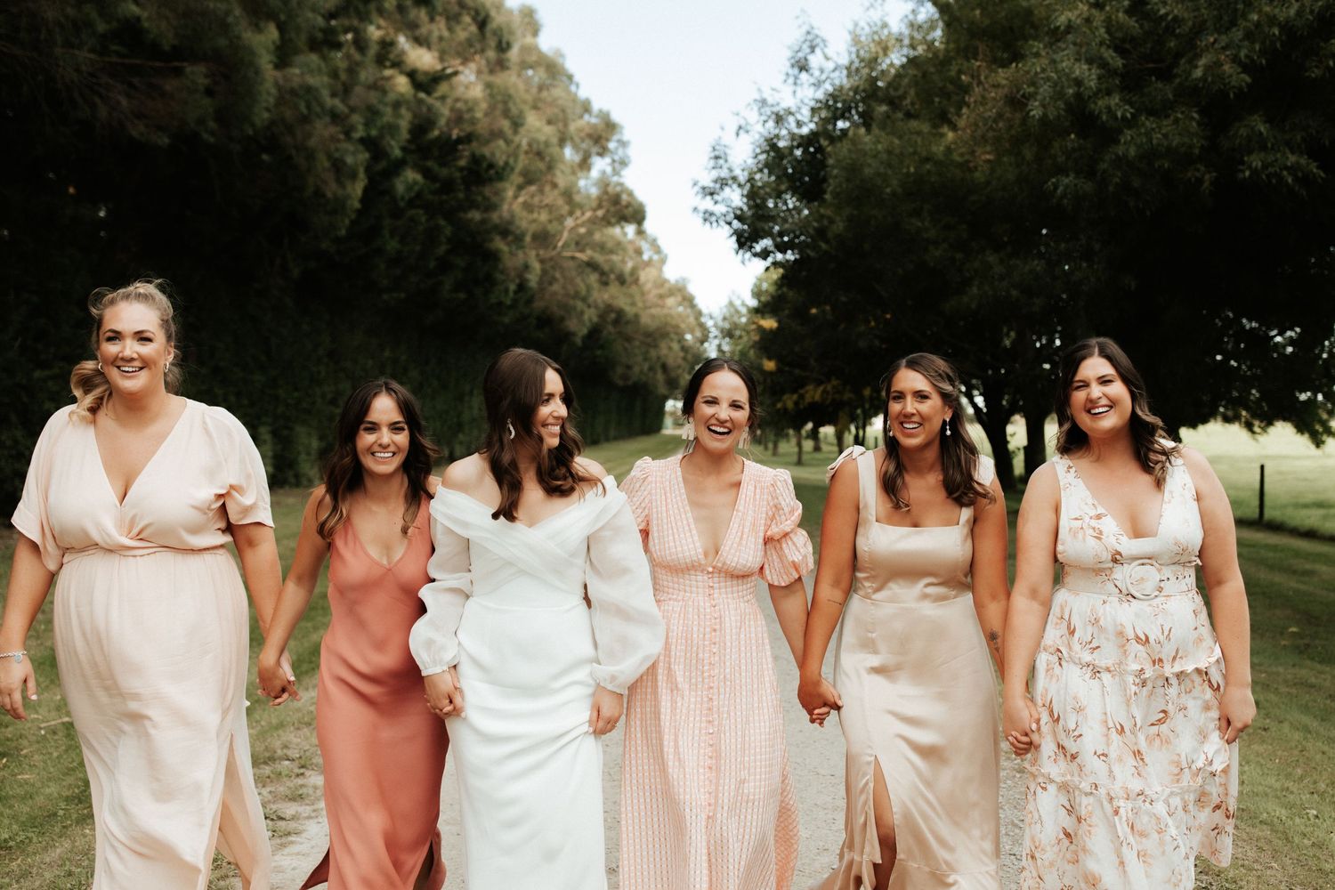 A group of bridesmaids in pastel dresses walk together on a scenic outdoor path.
