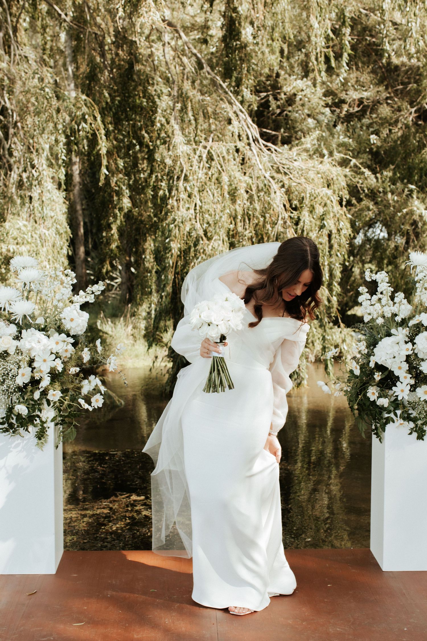 A bride in an elegant long-sleeved white dress holds her bouquet against a romantic outdoor backdrop.