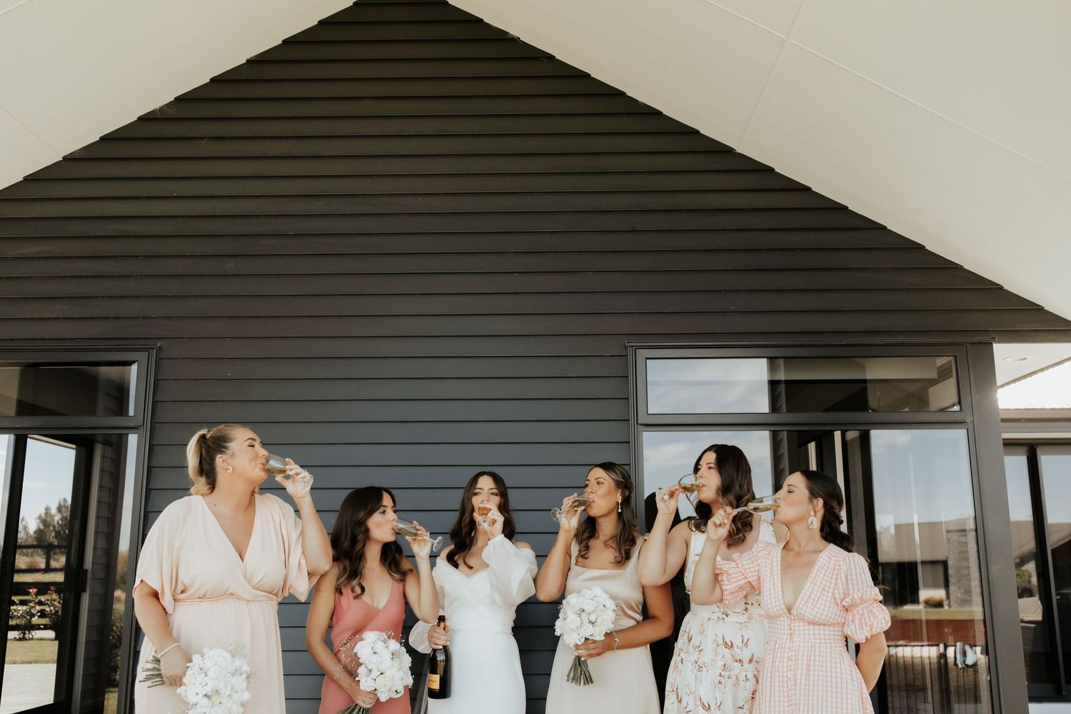 Bridesmaids in light-colored dresses gather for photos outside a modern house.