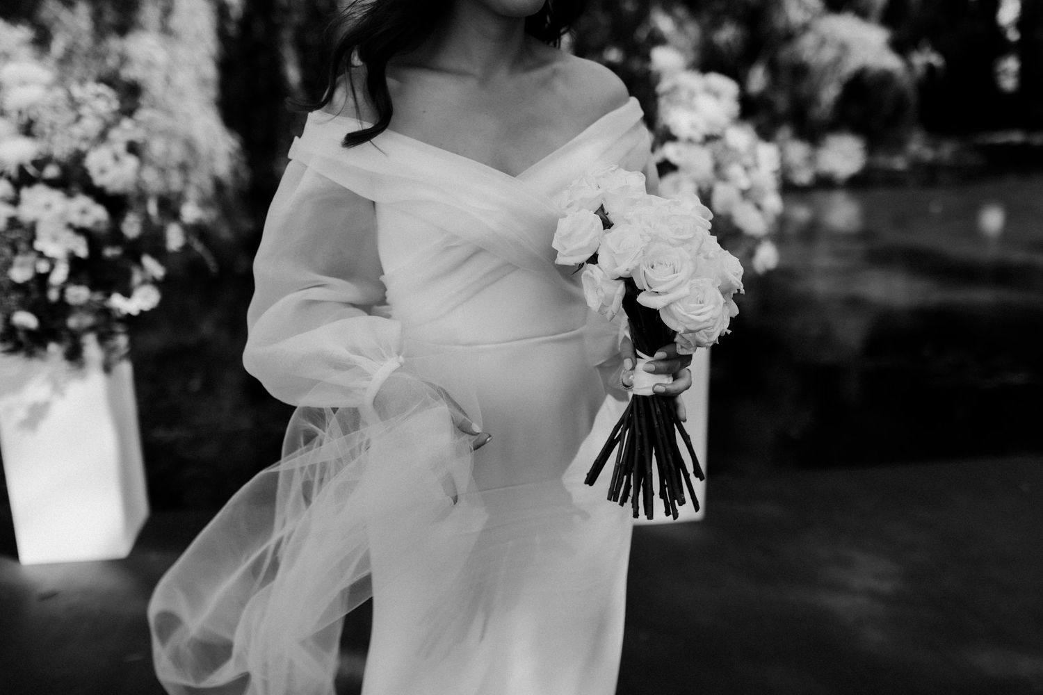 A black and white close-up of a bride in an elegant off-shoulder wedding dress holding a white floral bouquet.