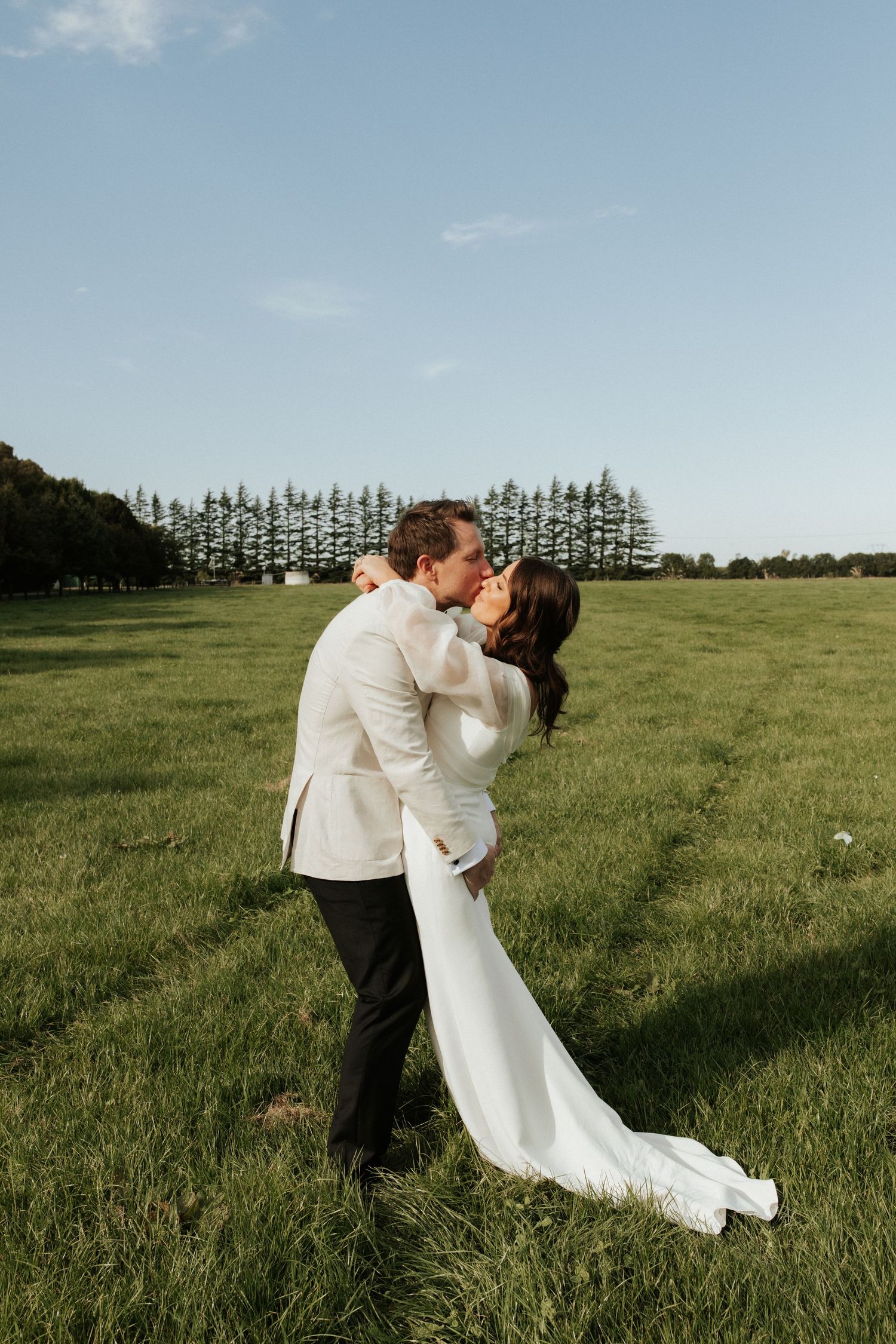 Romantic couple sharing a kiss in formal attire against a backdrop of pine trees.
