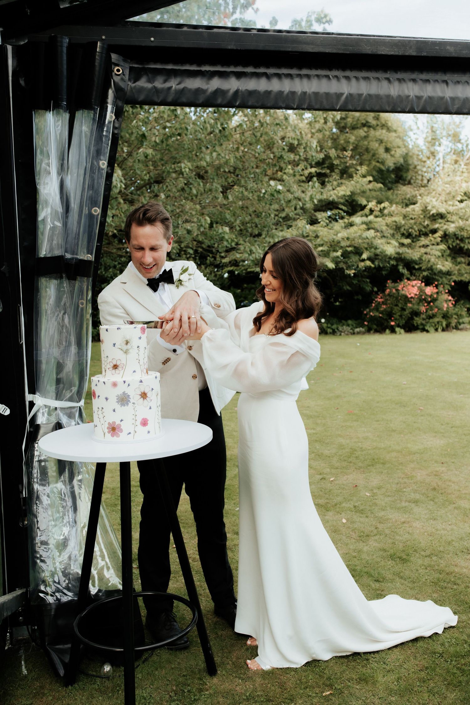 Couple cuts their floral-decorated wedding cake together under an outdoor pavilion.