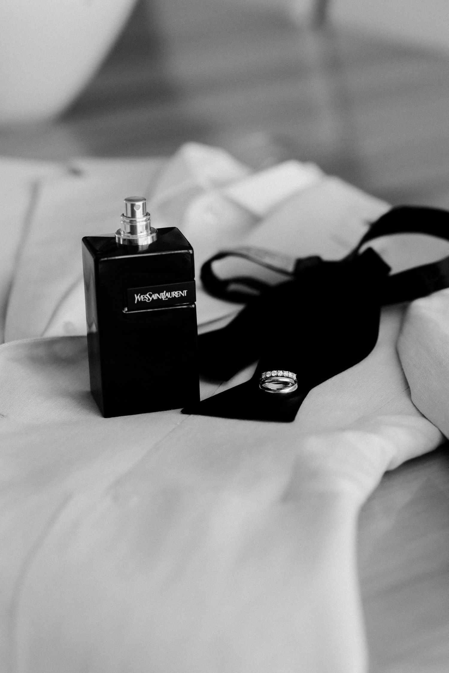 Black perfume bottle and strap displayed on white bedding in black and white.
