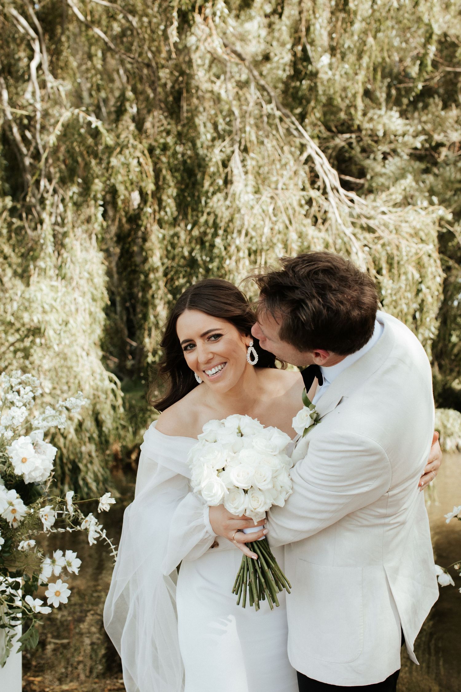 A romantic wedding moment captured under a beautiful willow tree with white floral arrangements.