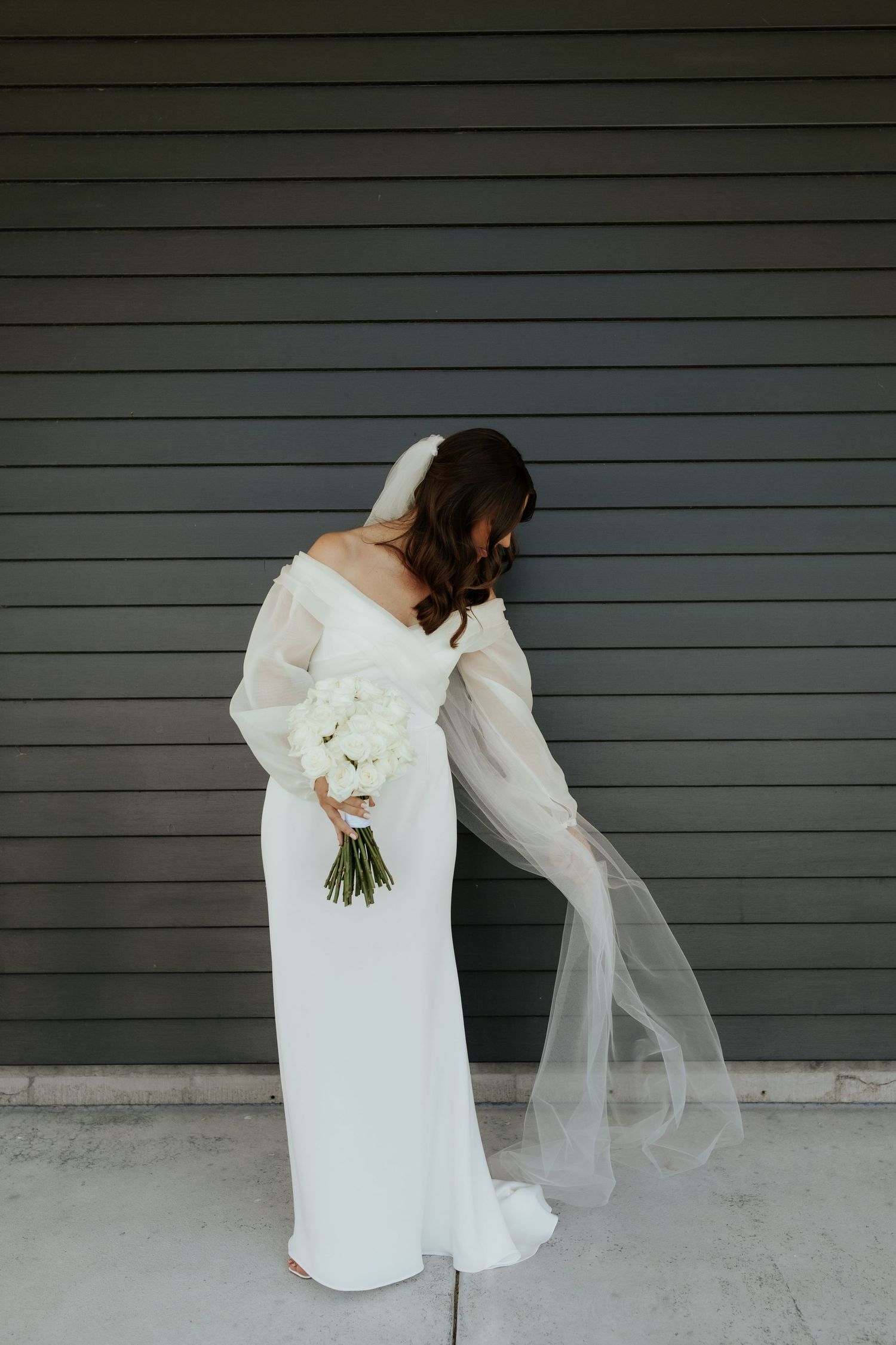 Bride in elegant white gown holding bouquet against gray wall.